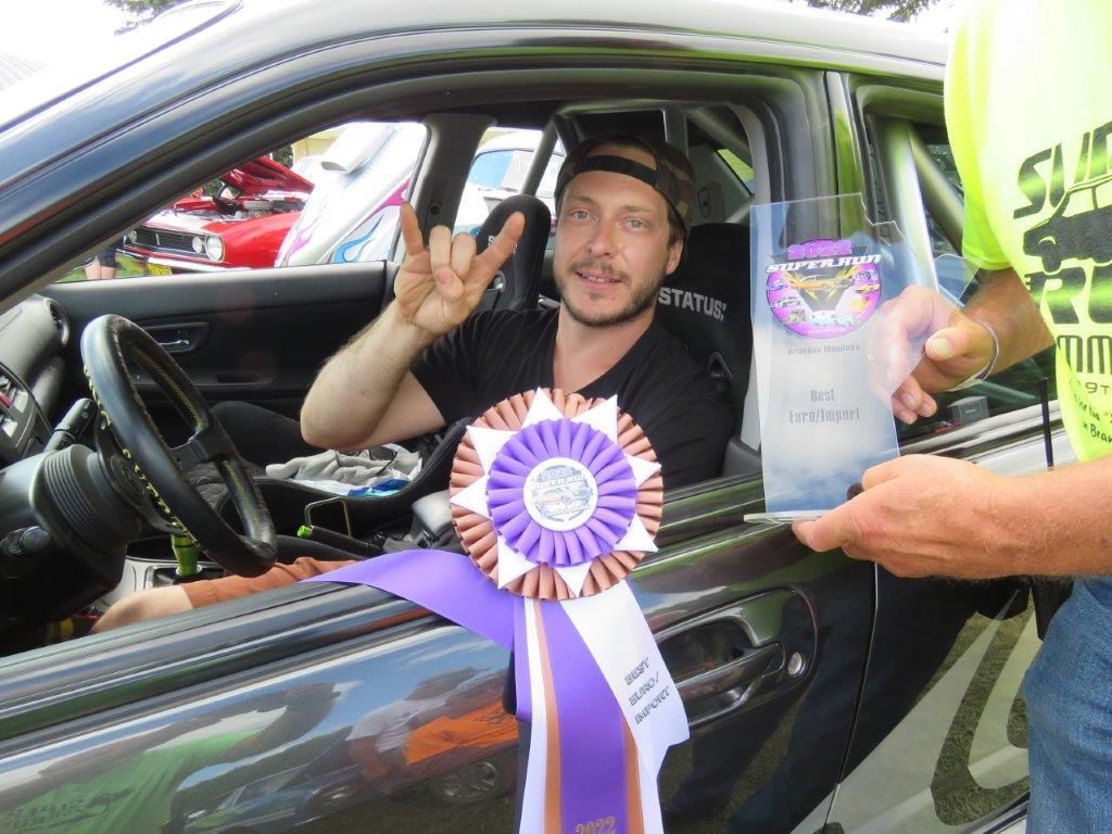 A man in a car with a purple ribbon and a trophy
