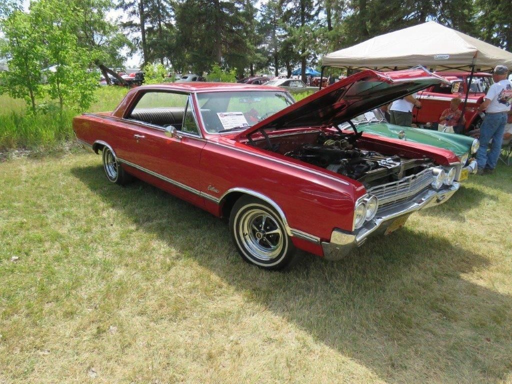 A red car with the hood up is parked in a grassy field.