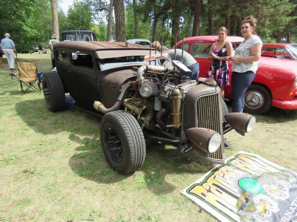 Two women are standing next to a rusty old car at a car show.
