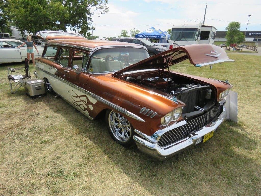 A brown and white car with the hood up is parked in a grassy field.
