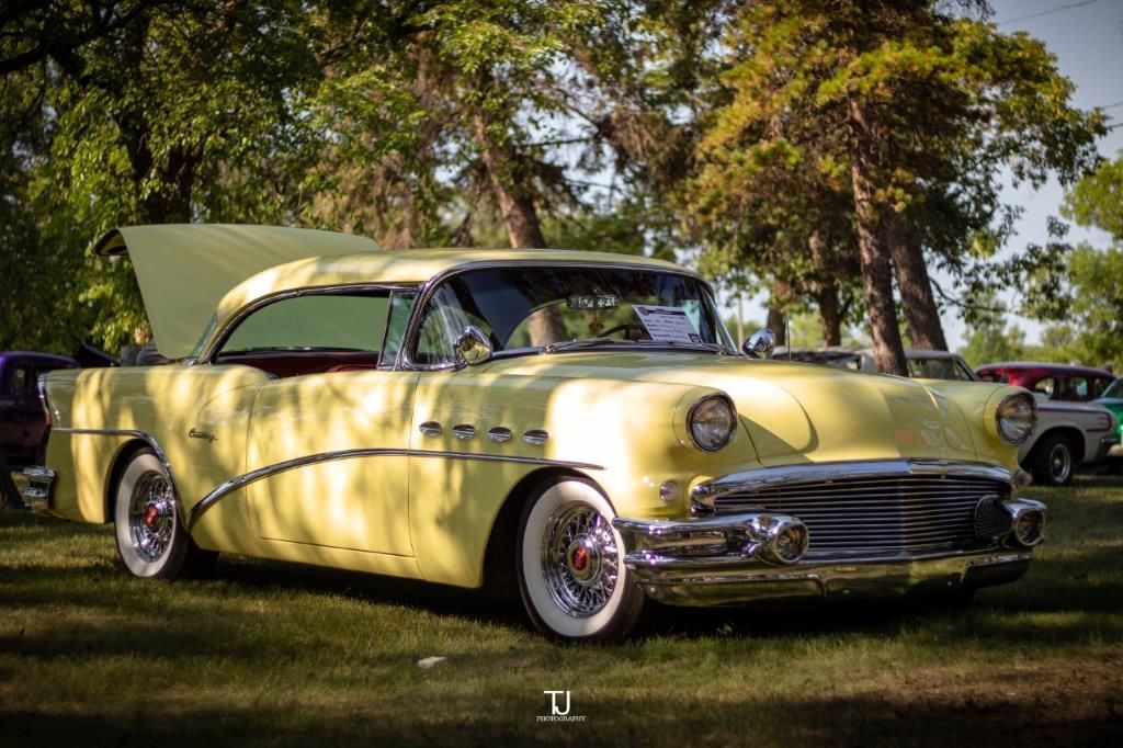 An old yellow car with the hood up is parked in the grass.
