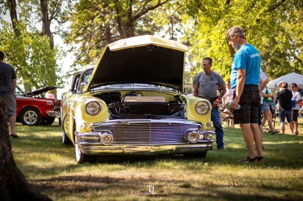 A man is standing in front of a yellow car with the hood up at a car show.
