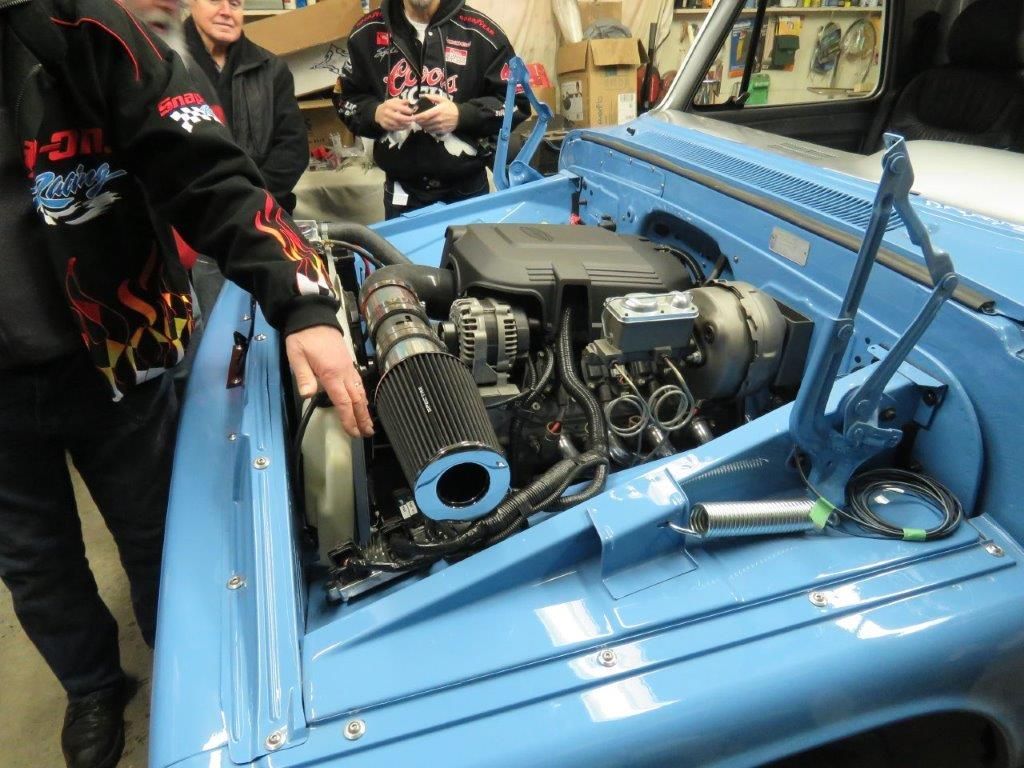 A group of men are looking under the hood of a blue truck.