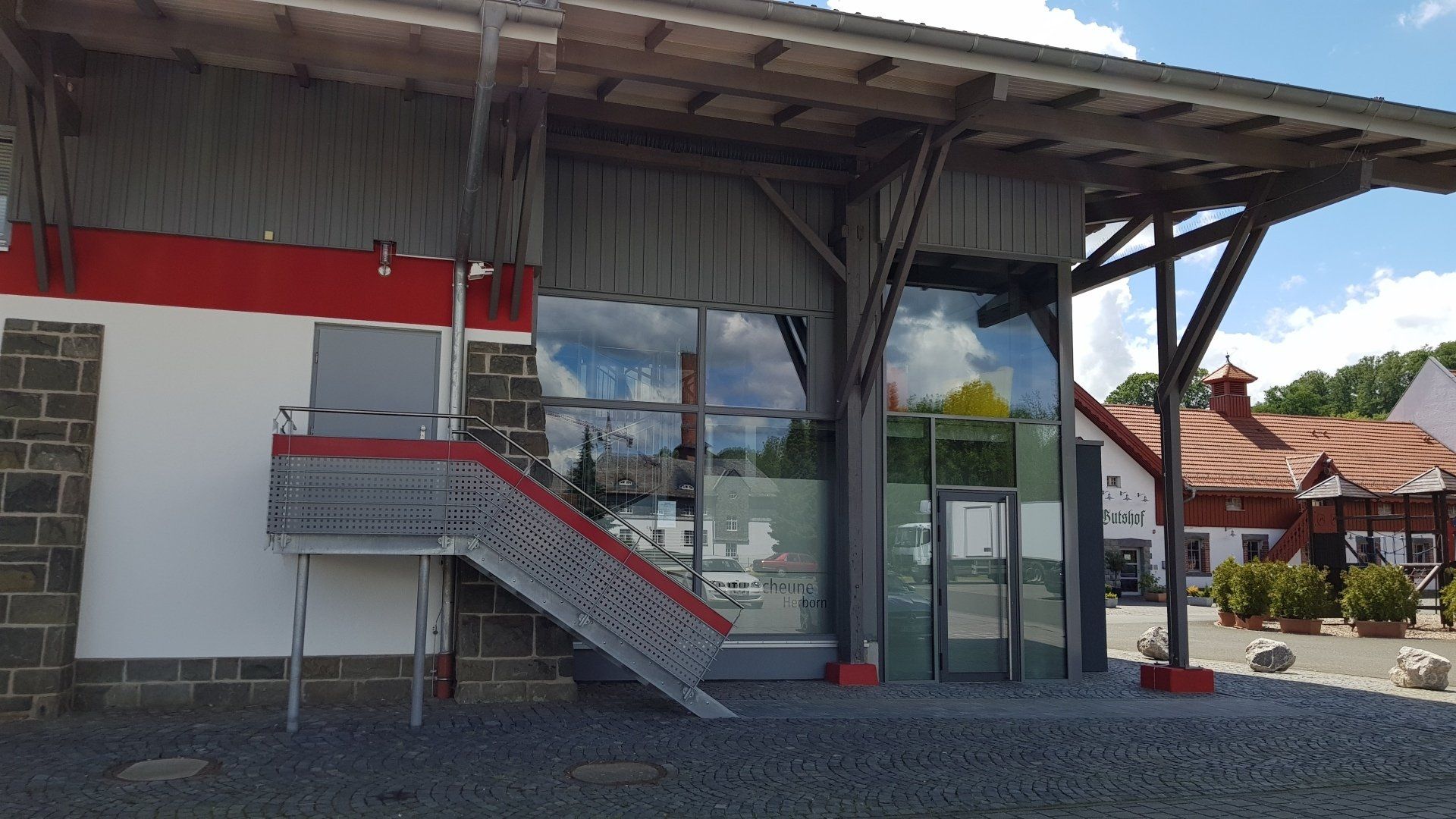 Moderner Gebäudeeingang mit Metalltreppe, großen Glasfenstern und Steinelementen vor einem Hintergrund aus Kiesfassade.