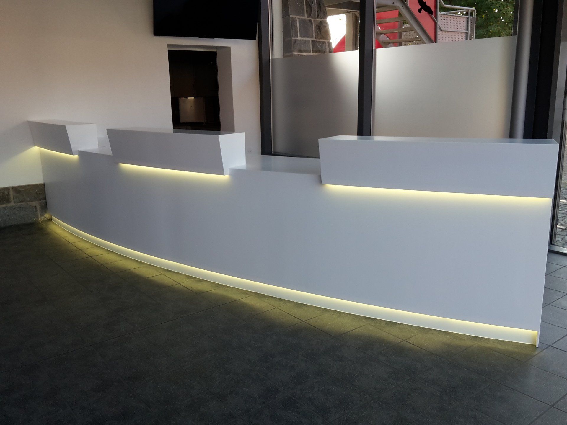 Ein weißer Empfangstresen mit integrierter warmweißer LED-Beleuchtung in einem Raum mit dunklem Fliesenboden.