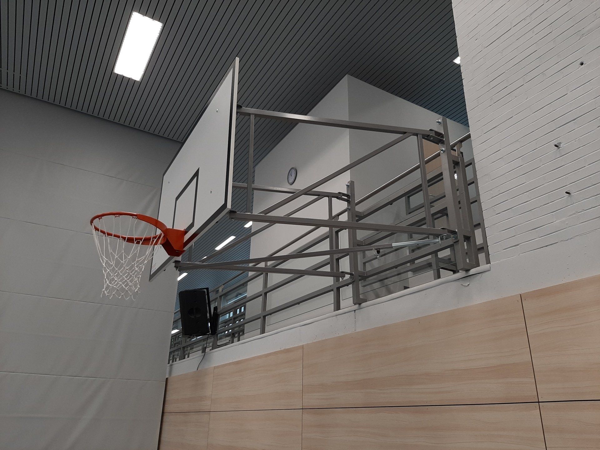 Ein Basketballkorb mit weißem Brett ist an einem klappbaren Metallgestell an einer Wand einer Sporthalle befestigt.