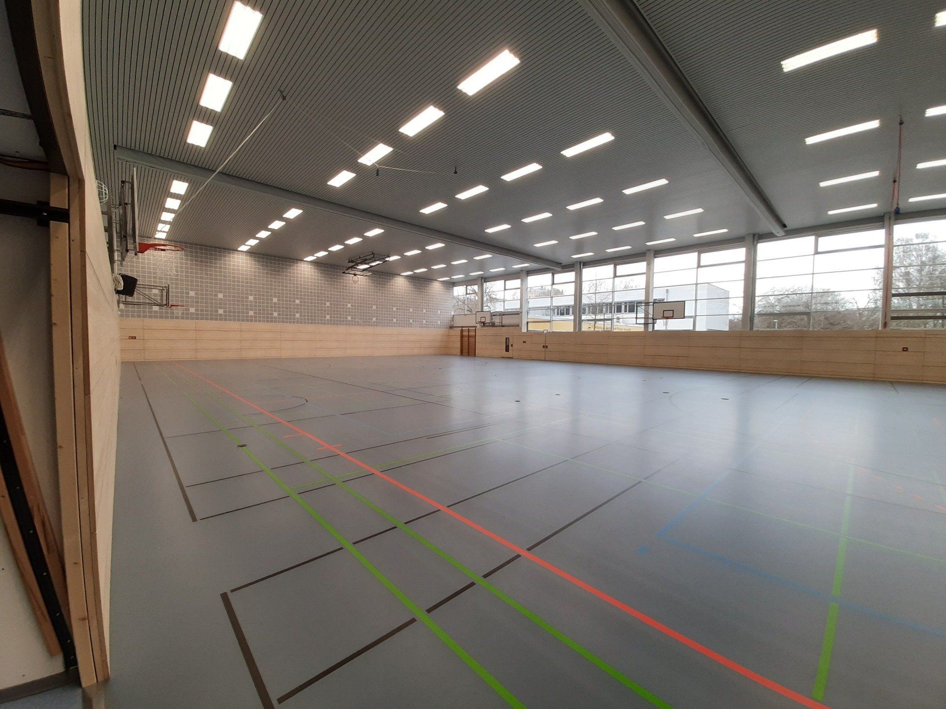 Eine leere Turnhalle mit grauem Boden, holzgetäfelten Wänden und Reihen von Deckenleuchten.