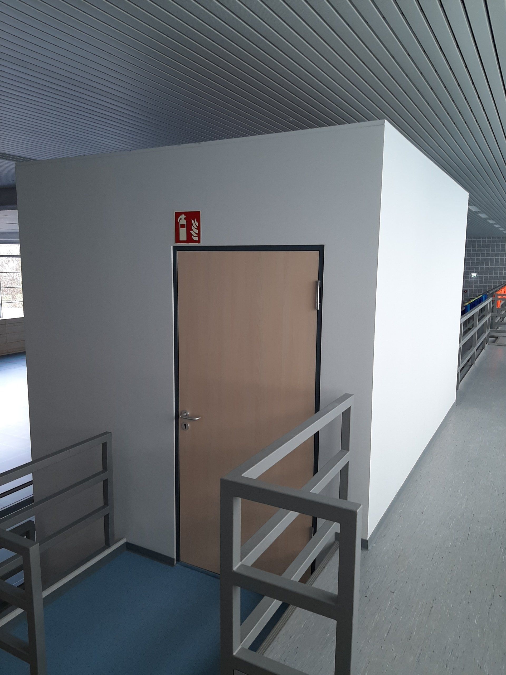 Eine hellbraune Tür in einer weißen Wand, darüber ein rotes Feuerlöscher-Schild und im Vordergrund ein Metallgeländer.