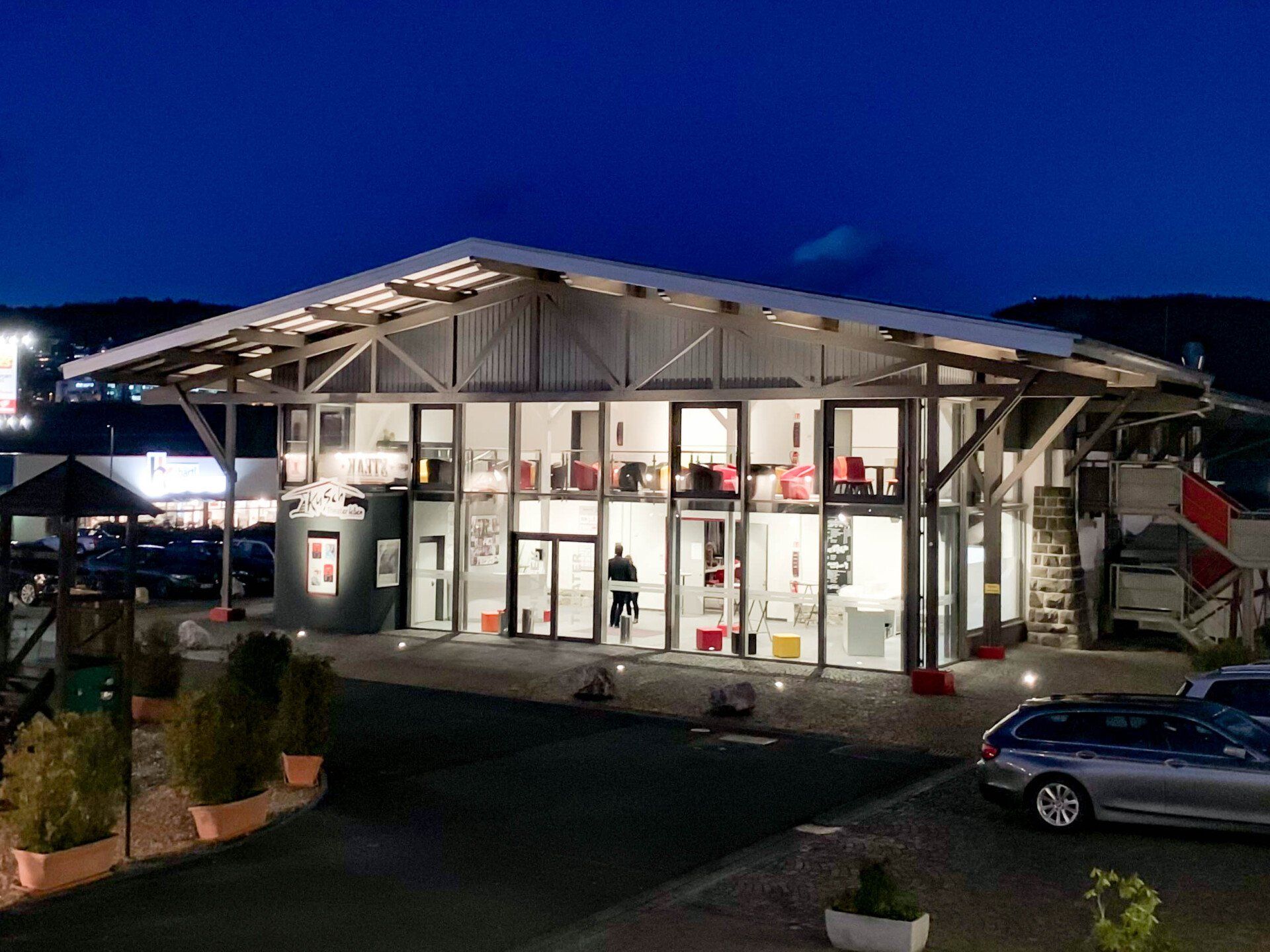 Modernes zweistöckiges Gebäude mit Glasfassade bei Nacht, mit Satteldach, Außenbeleuchtung und in der Nähe geparkten Autos.