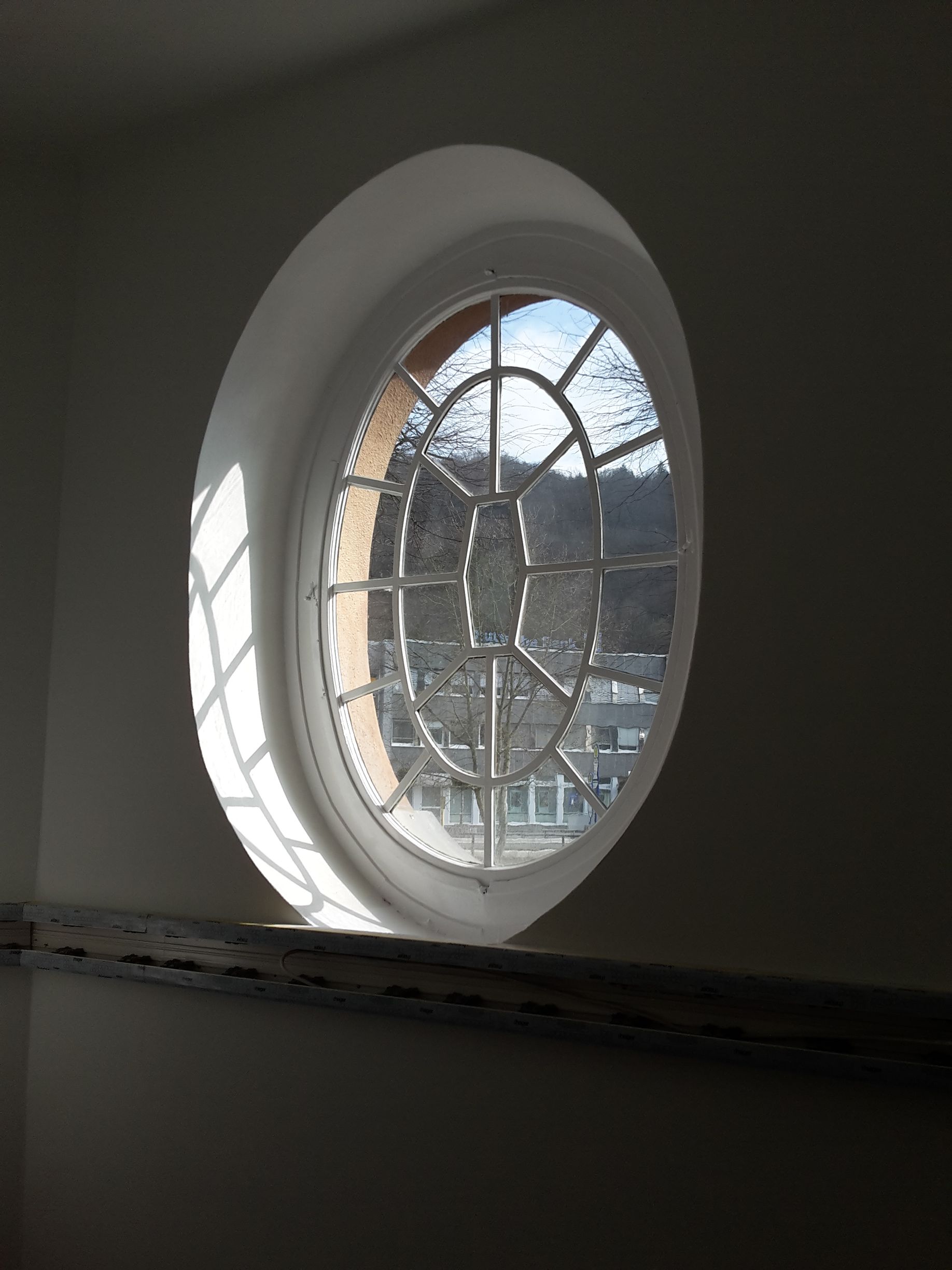 Ein ovales Fenster mit dekorativem Sprossenmuster, das von Sonnenlicht erhellt wird, welches einen gitterförmigen Schatten