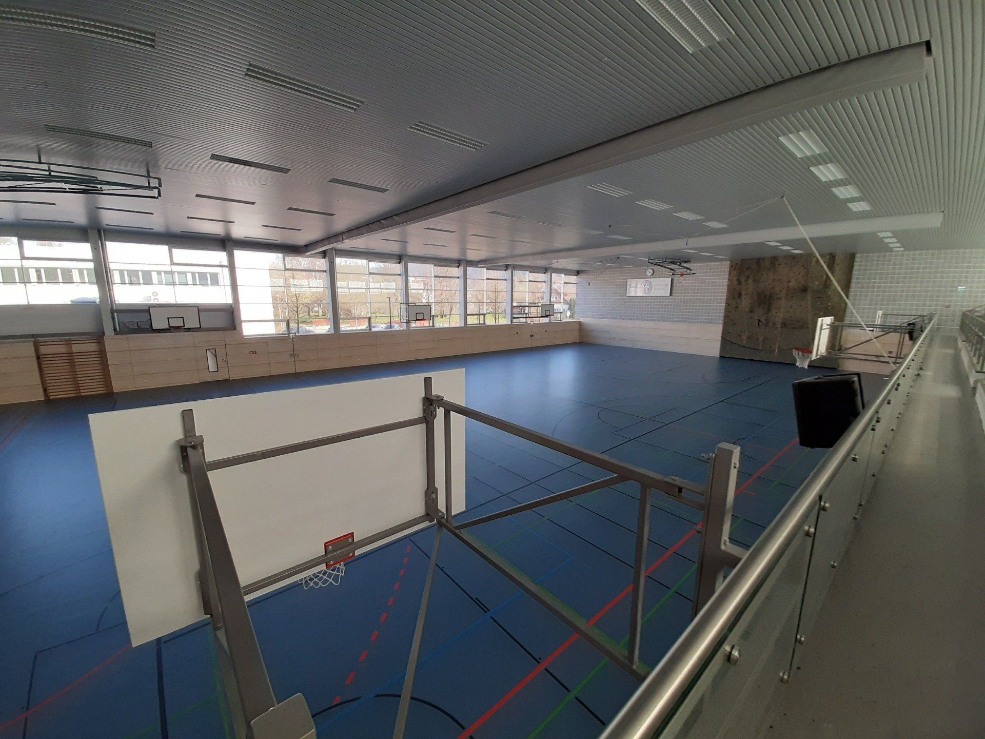 Eine Turnhalle mit blauem Boden, einem Basketballkorb im Vordergrund, einer Kletterwand und großen Fenstern.