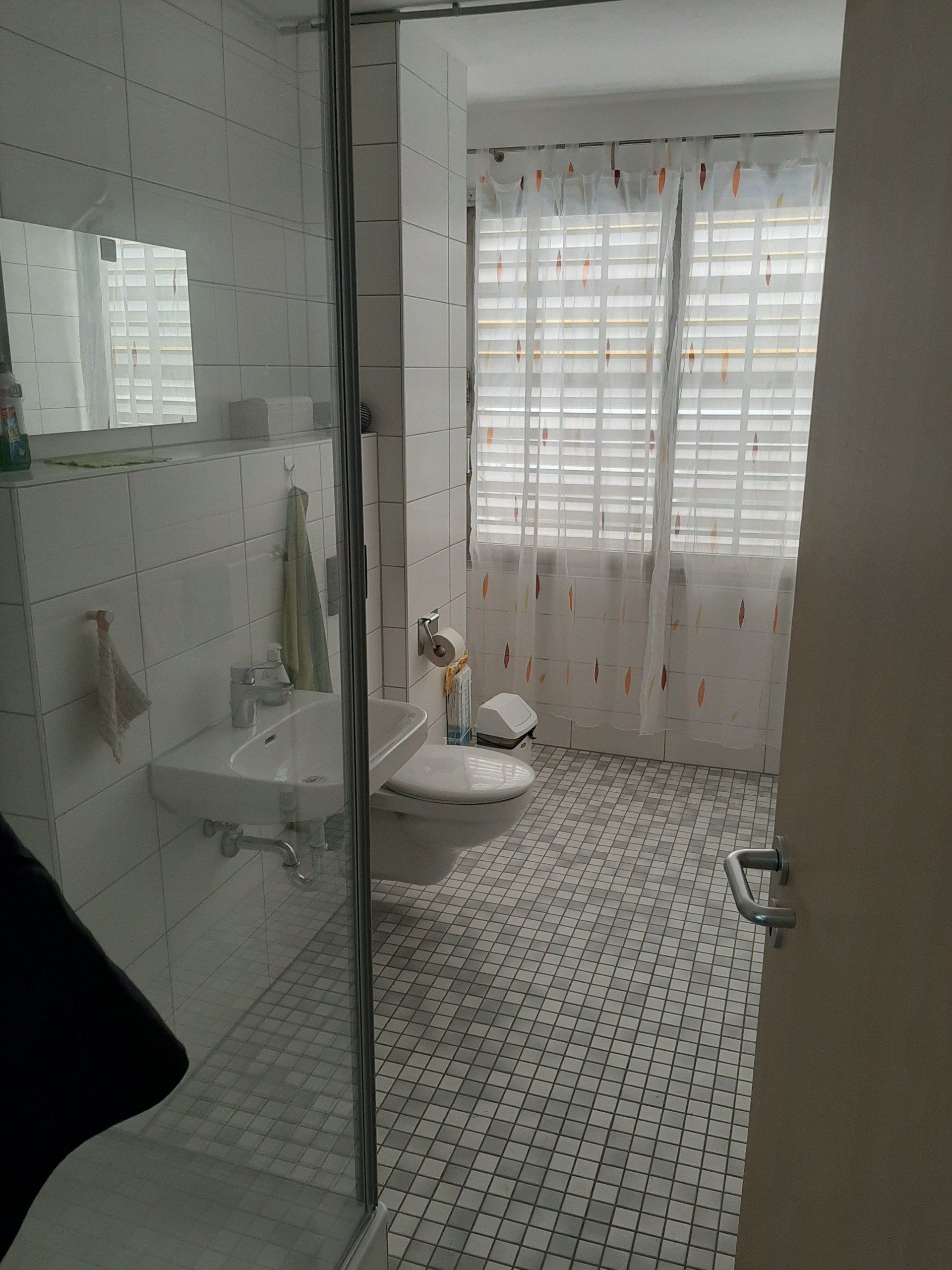 Ein weiß gefliestes Badezimmer mit Waschbecken, Toilette, Duschkabine und Fenster mit transparenten Vorhängen.