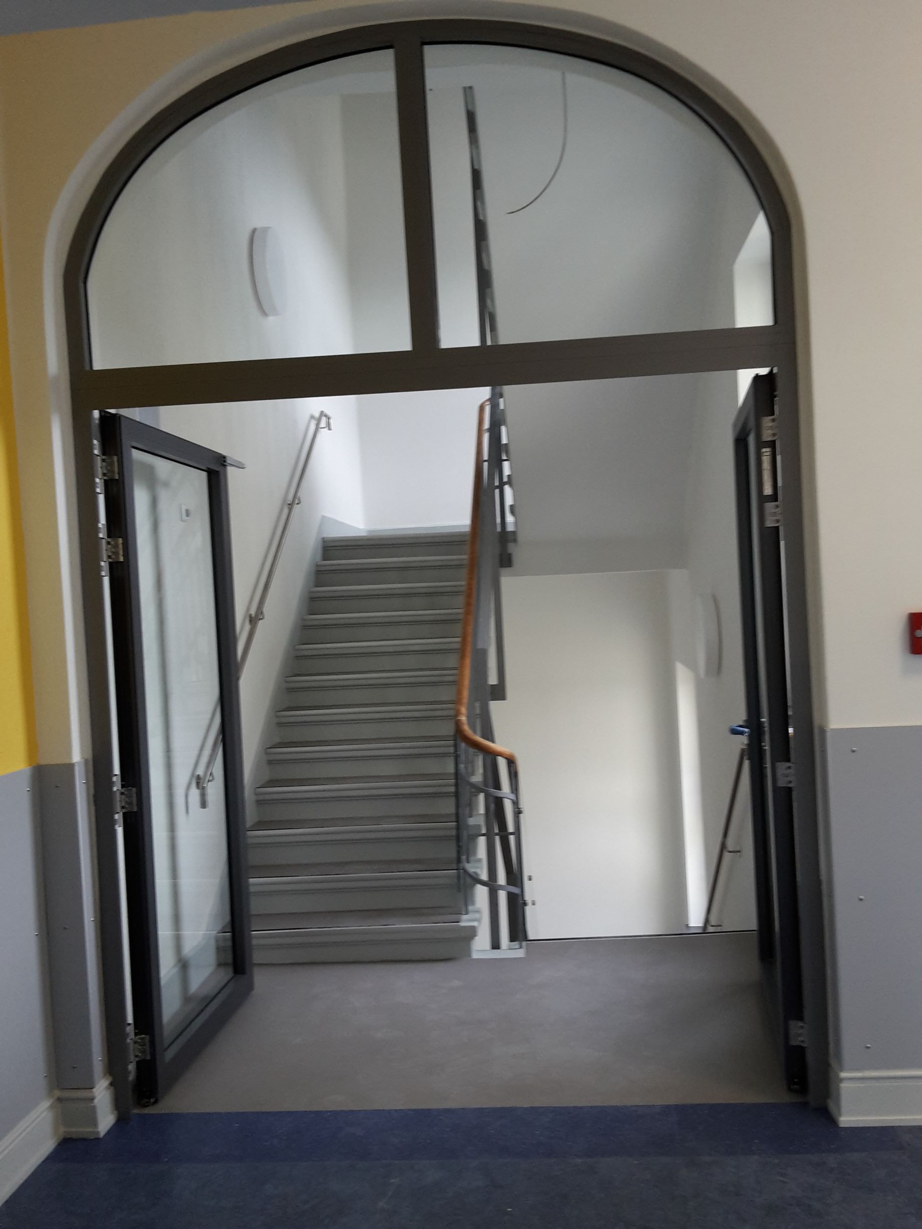 Der Blick durch einen Bogengang in ein Treppenhaus mit grauer Treppe, hölzernem Handlauf und weißen Wänden.