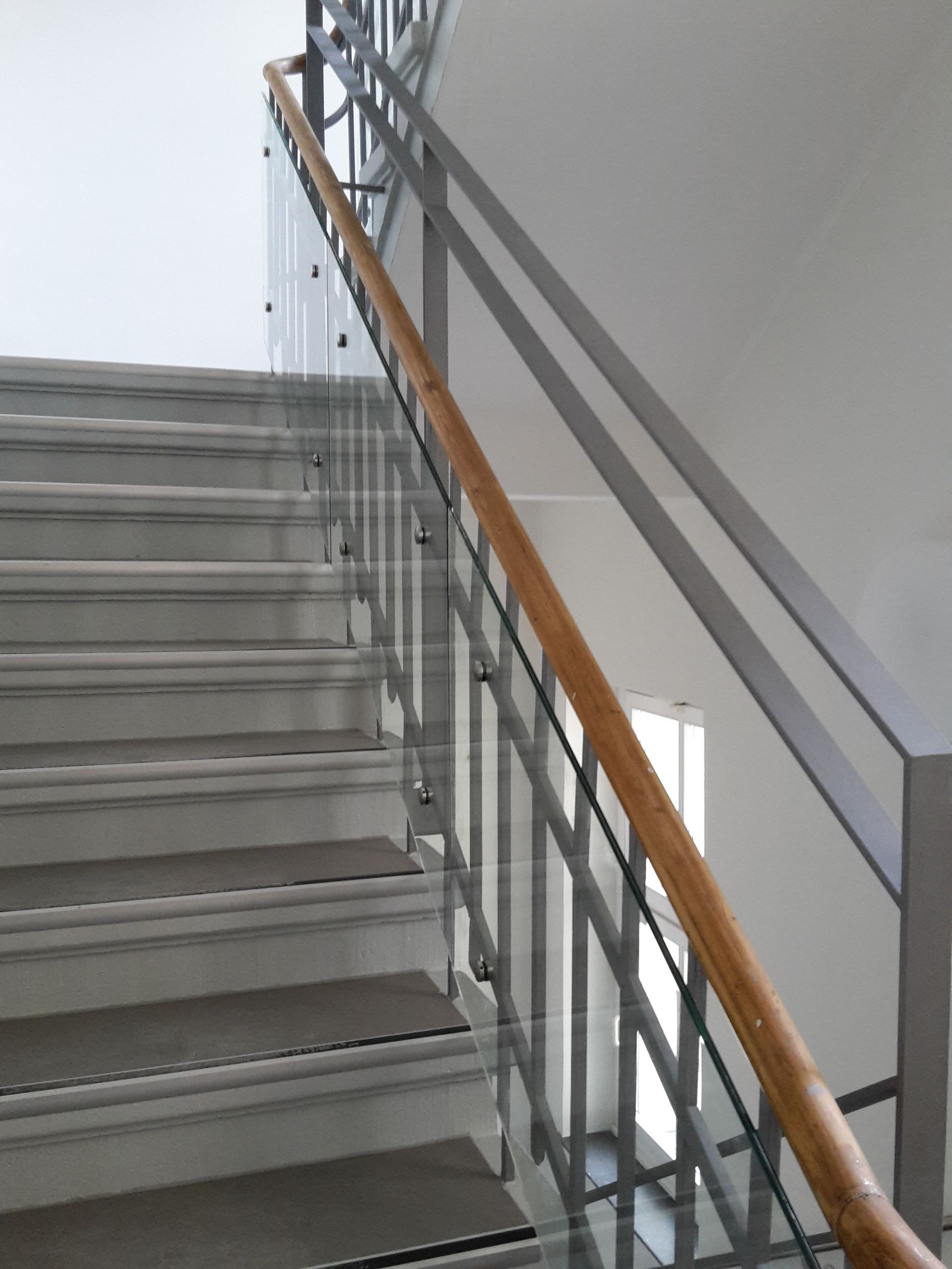 Eine Treppe mit Betonstufen mit grauen Trittflächen, einem Glasgeländer und einem warmfarbigen Holzhandlauf.