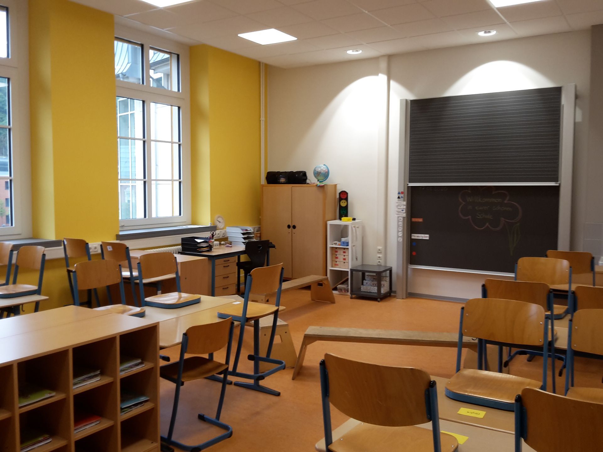 Leeres Klassenzimmer mit hölzernen Schülerstühlen, Schreibtischen, Regalen und einer Tafel vor einer gelb-weißen Wand.