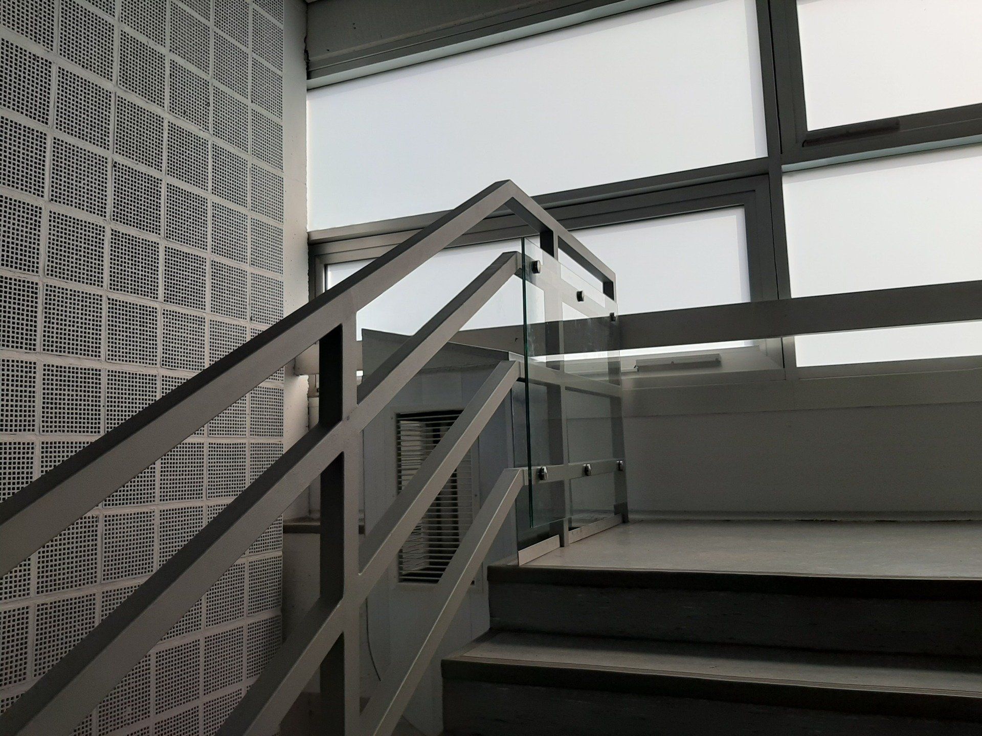 Eine Treppe mit Metallgeländer und Glasscheibe führt hinauf zu einem hellen Fenster neben einer gemusterten weißen Wand.