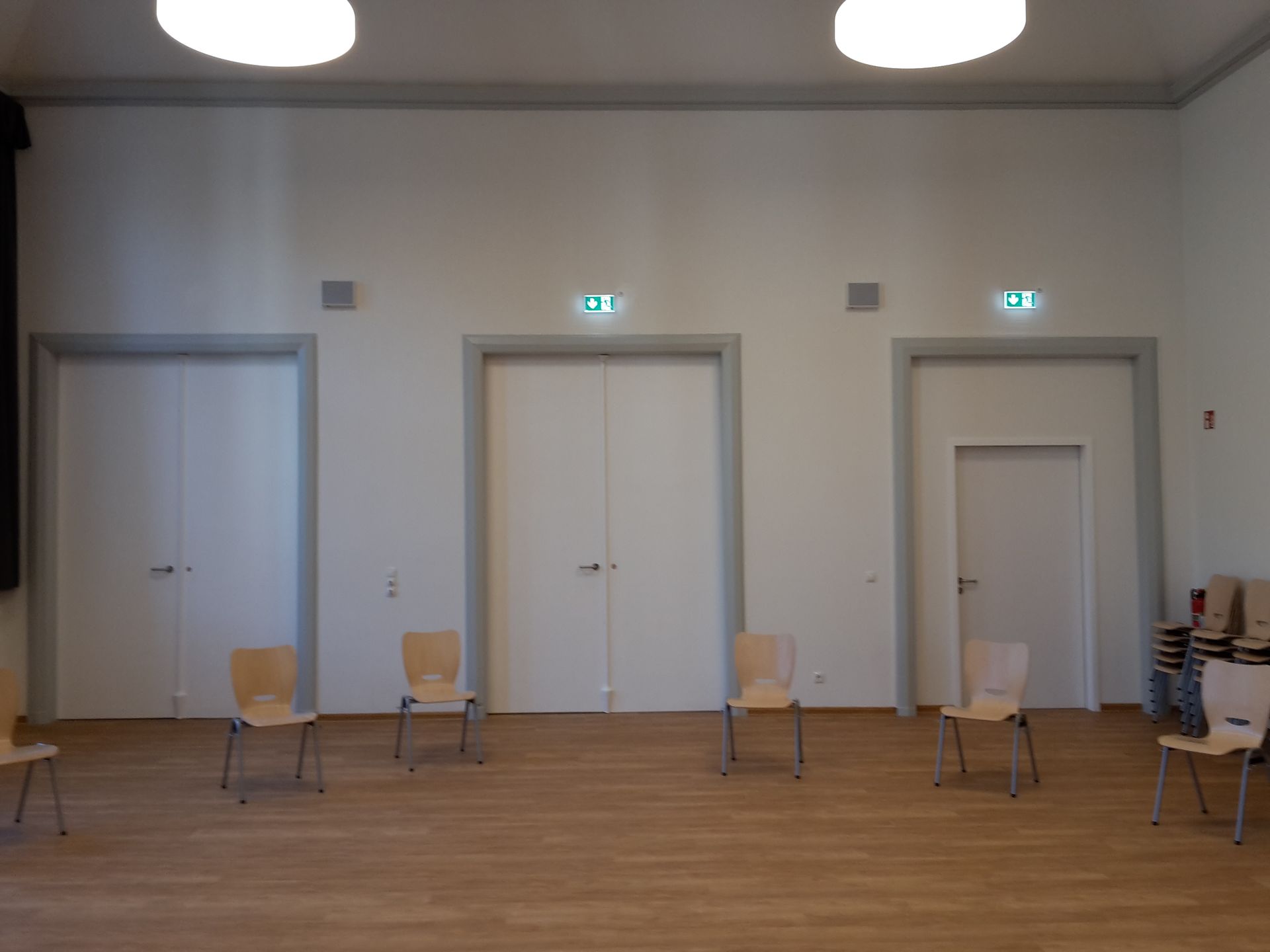Ein Zimmer mit Holzfußboden, weißen Wänden, drei Türrahmen und fünf leeren Stühlen, die in einer Reihe aufgestellt sind.