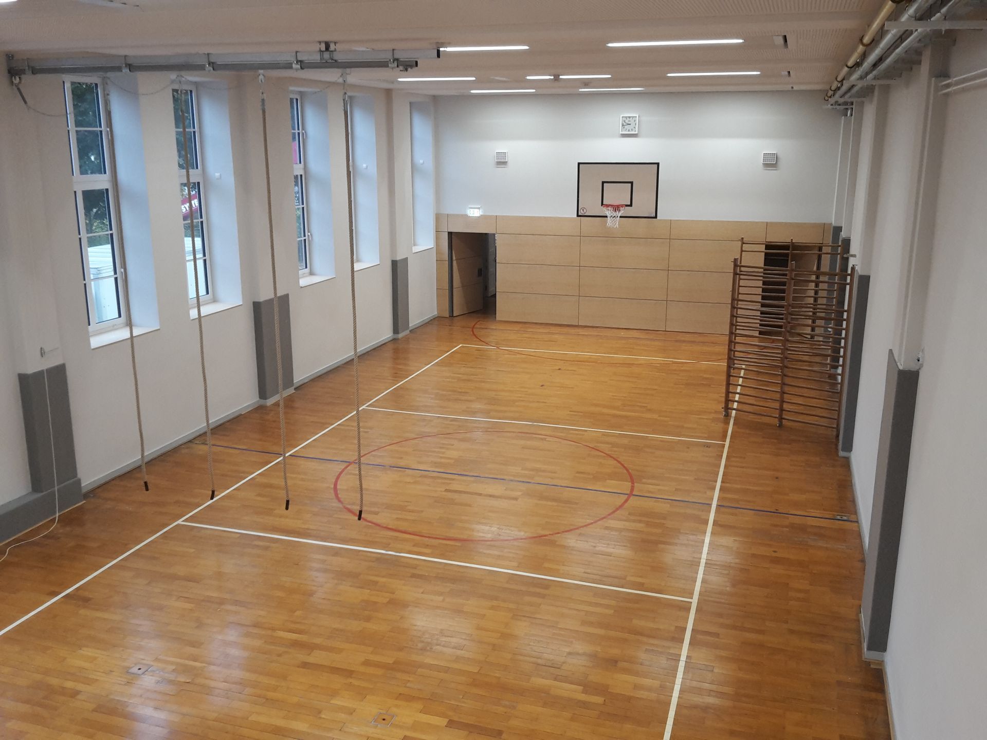 Eine leere Turnhalle mit Holzboden, Basketballkorb, weißen Wänden, großen Fenstern und einer Sprossenwand.