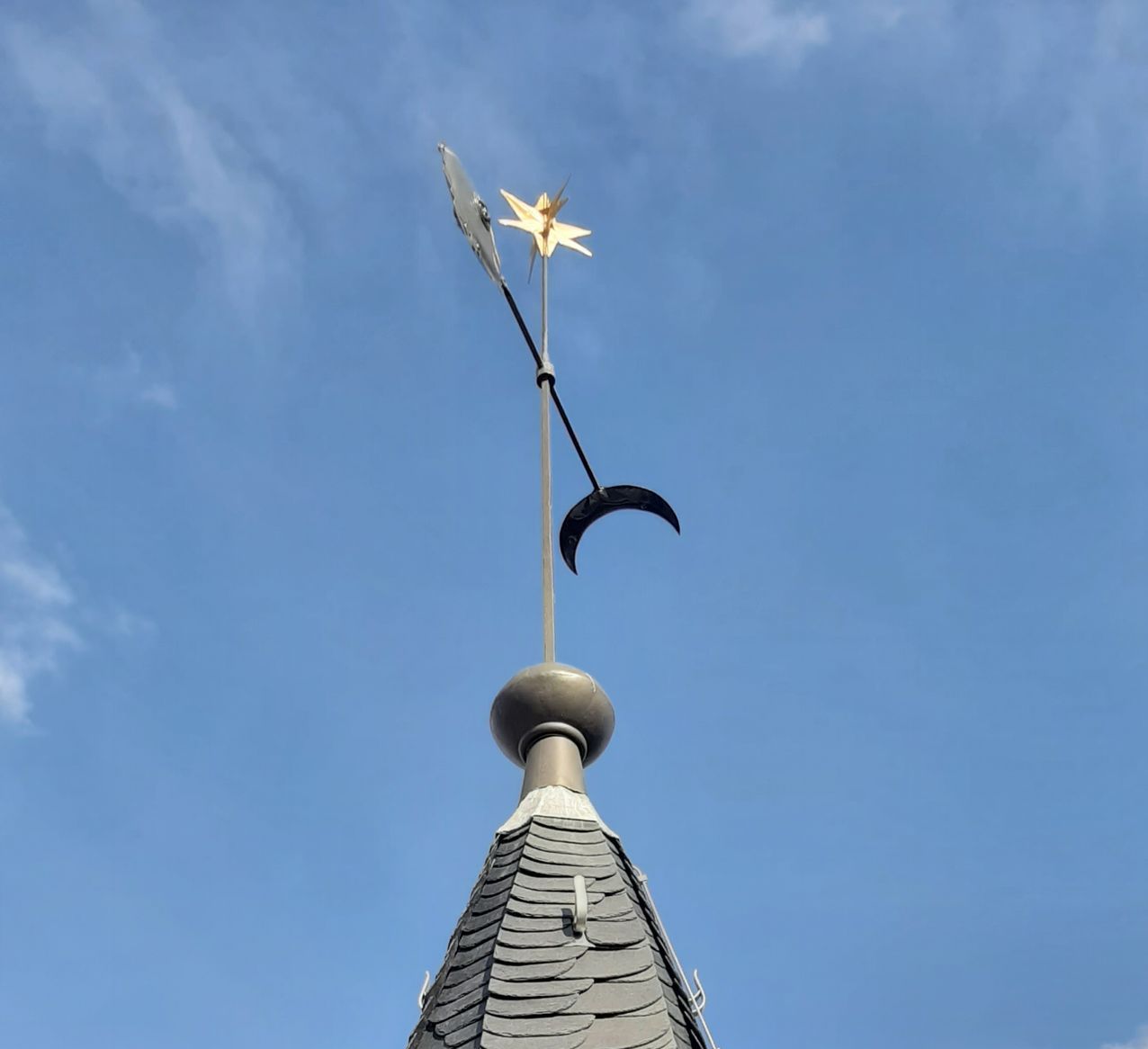 Ein goldener Stern und eine schwarze, halbmondförmige Metallfigur thronen auf einem Kirchturm vor einem klaren blauen Himmel.