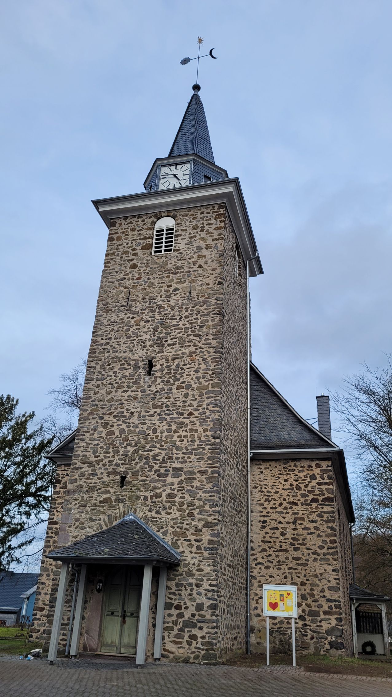 Steinkirche mit hohem Turm, spitz zulaufendem Schieferdach, Wetterfahne und Eingangshalle unter bewölktem Himmel.
