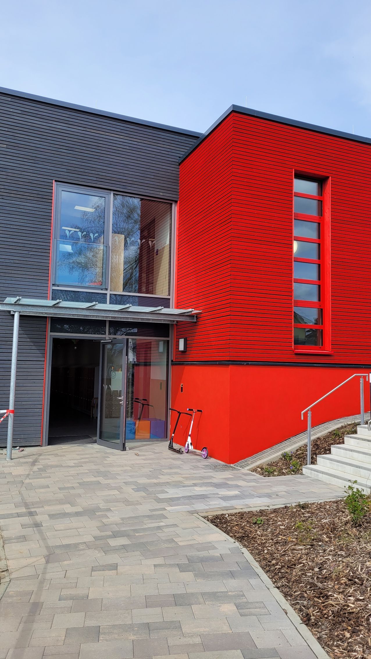 Moderner Gebäudeeingang mit kontrastierender roter Fassade, dunkler, strukturierter Verkleidung, großen Glastüren 