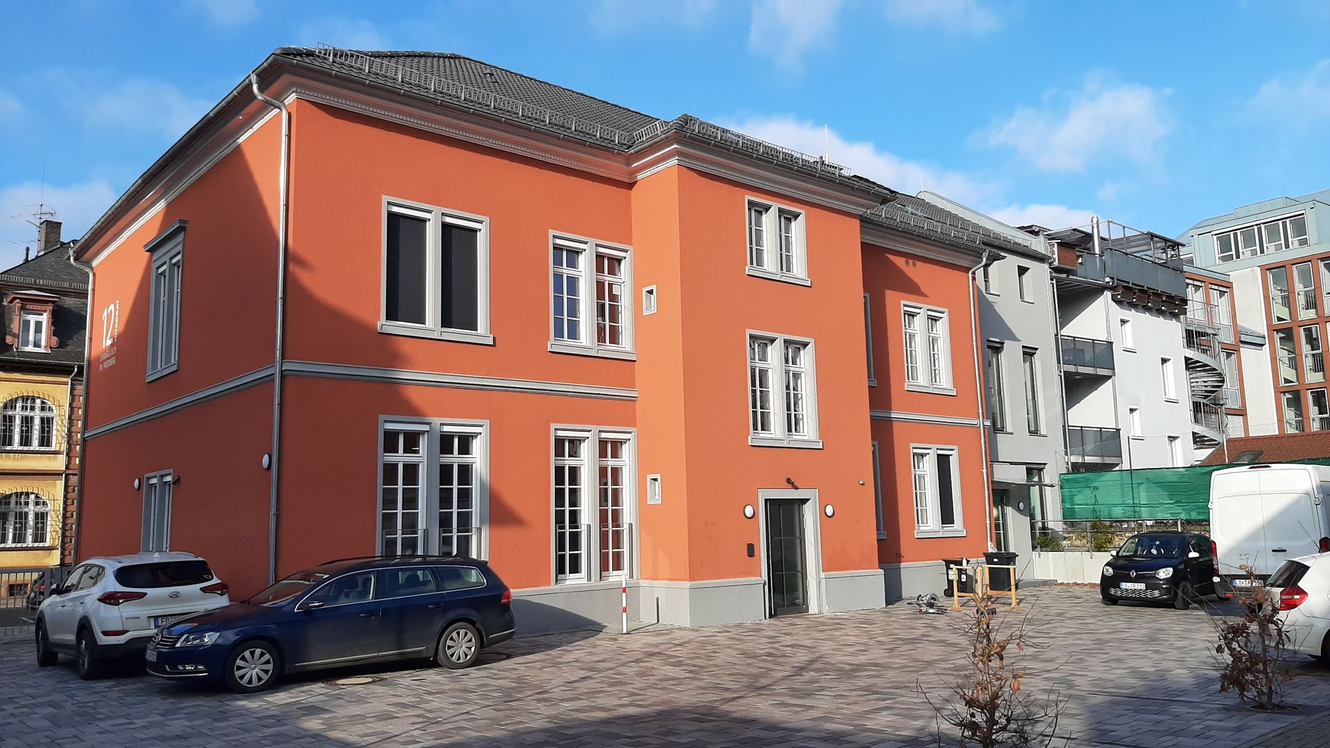 An einem sonnigen Tag steht in einem gepflasterten Innenhof mit geparkten Autos 
