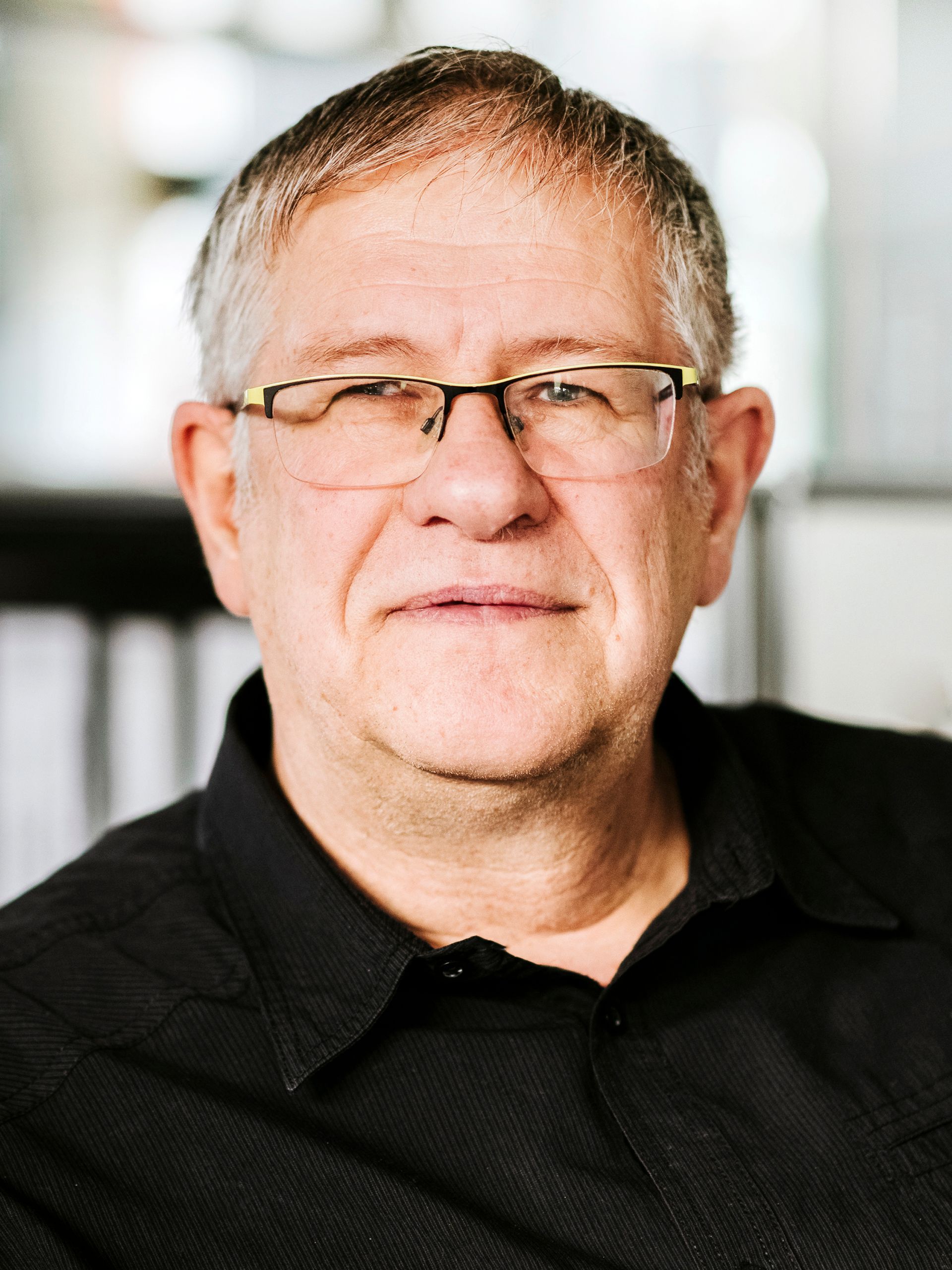 Ein Porträt einer Person mit Brille und schwarzem Hemdkragen, die vor verschwommenem Hintergrund in die Kamera blickt.