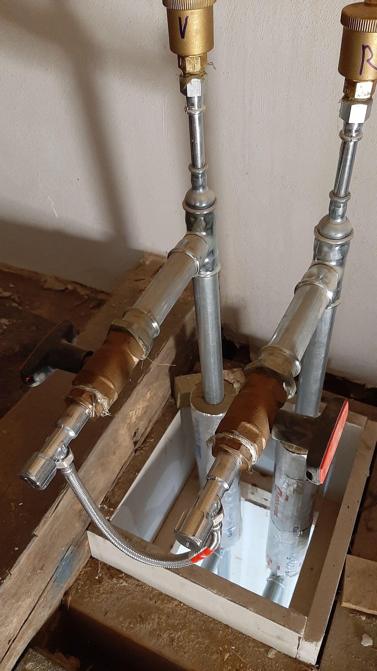 Zwei senkrechte Metallrohre mit Messingarmaturen ragen aus einer quadratischen Öffnung in einem Holzboden.