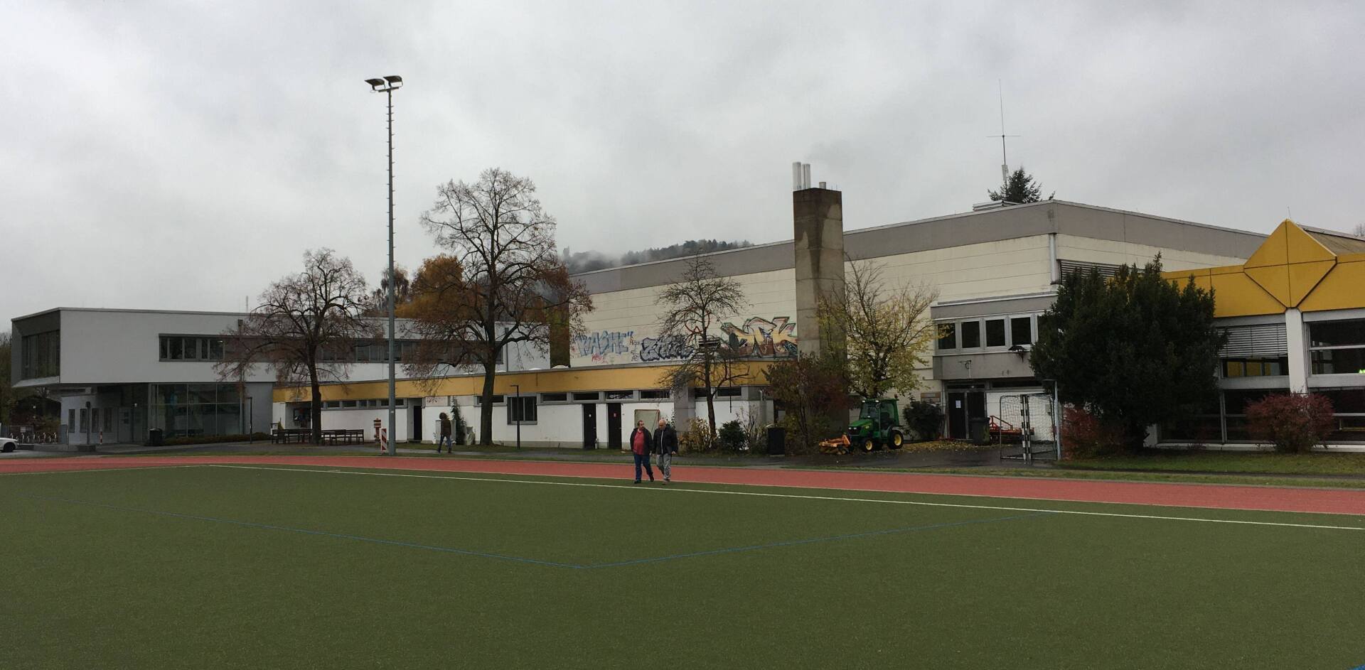Ein flaches, modernes Schulgebäude mit einer großen Turnhalle, gegenüber einem Kunstrasen-Sportplatz unter grauem Himmel.