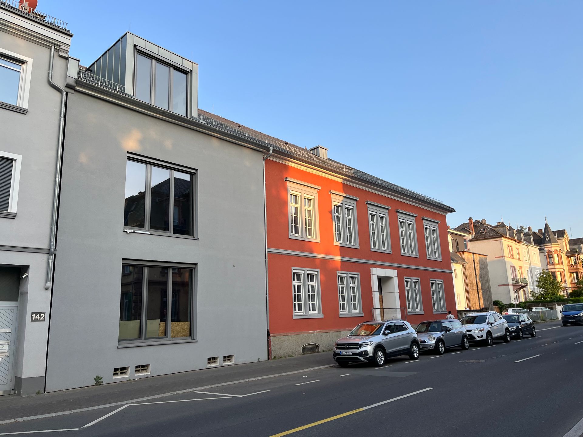 Blick von der Straße auf ein modernes, hellgraues Gebäude neben einem klassischen orangefarbenen Gebäude