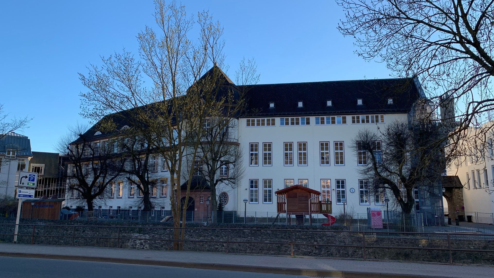 Ein weißes, zweistöckiges historisches Gebäude mit dunklem Schindeldach, umgeben von kahlen Bäumen unter klarem, 