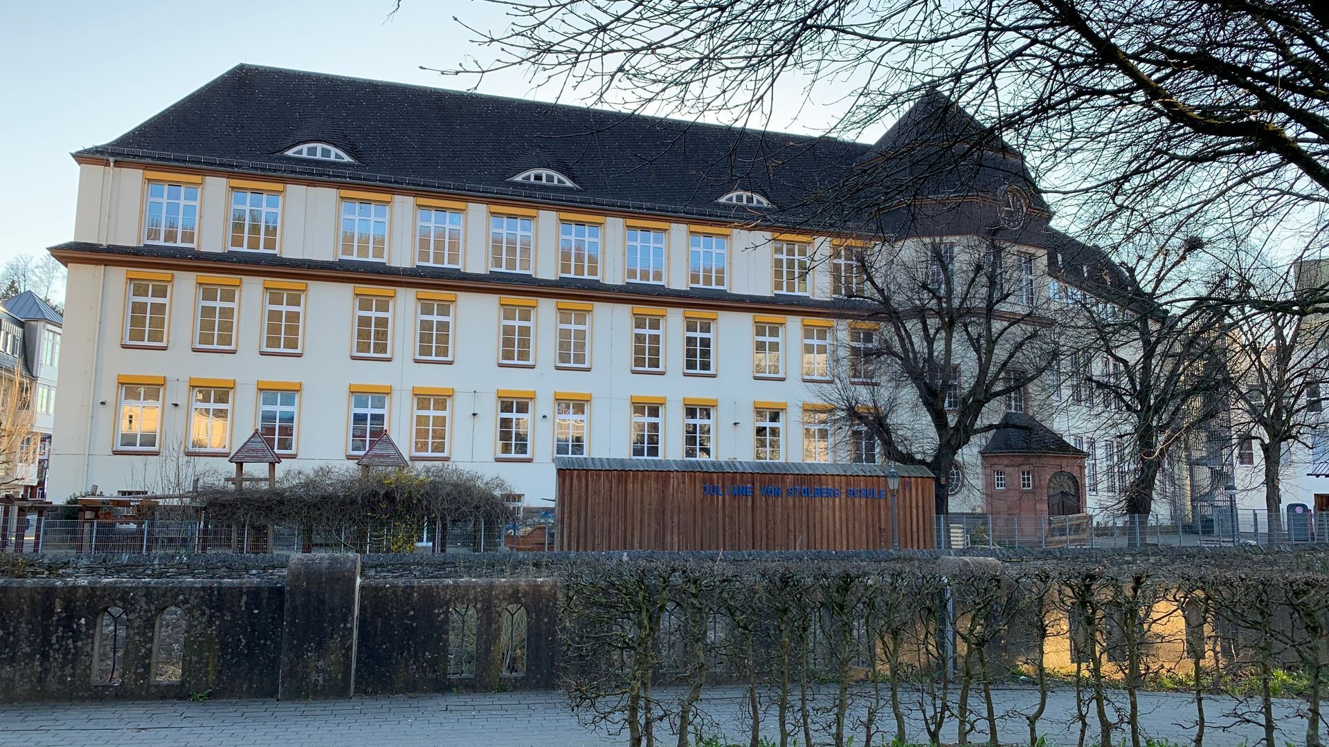 Hinter einer niedrigen Hecke und einem Holzzaun steht ein dreistöckiges, hellfarbiges historisches Schulgebäude mit 