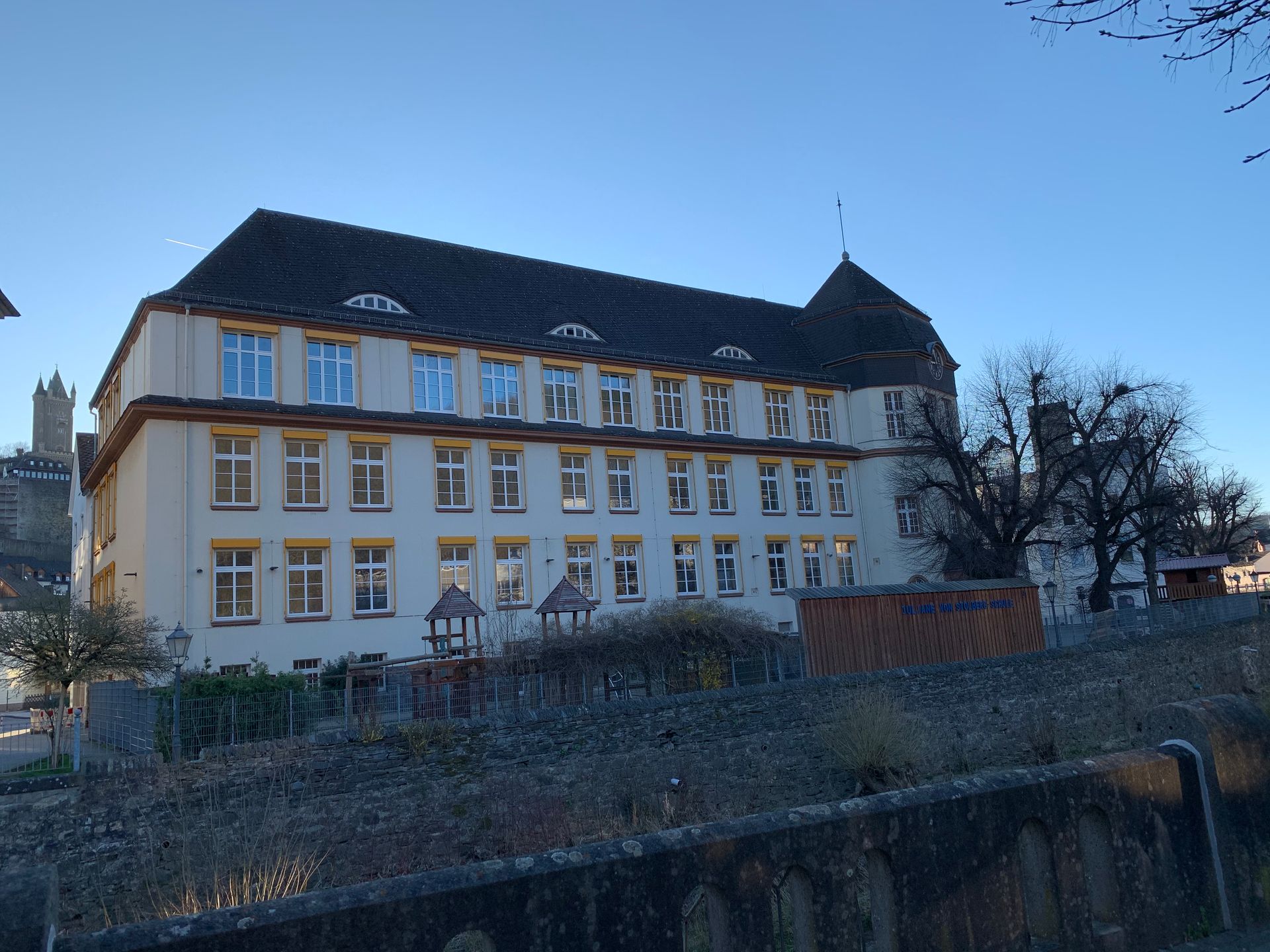 Ein großes, mehrstöckiges weißes Gebäude mit dunklem Dach und einem kleinen Turm, das sich hinter einer Steinmauer vor 