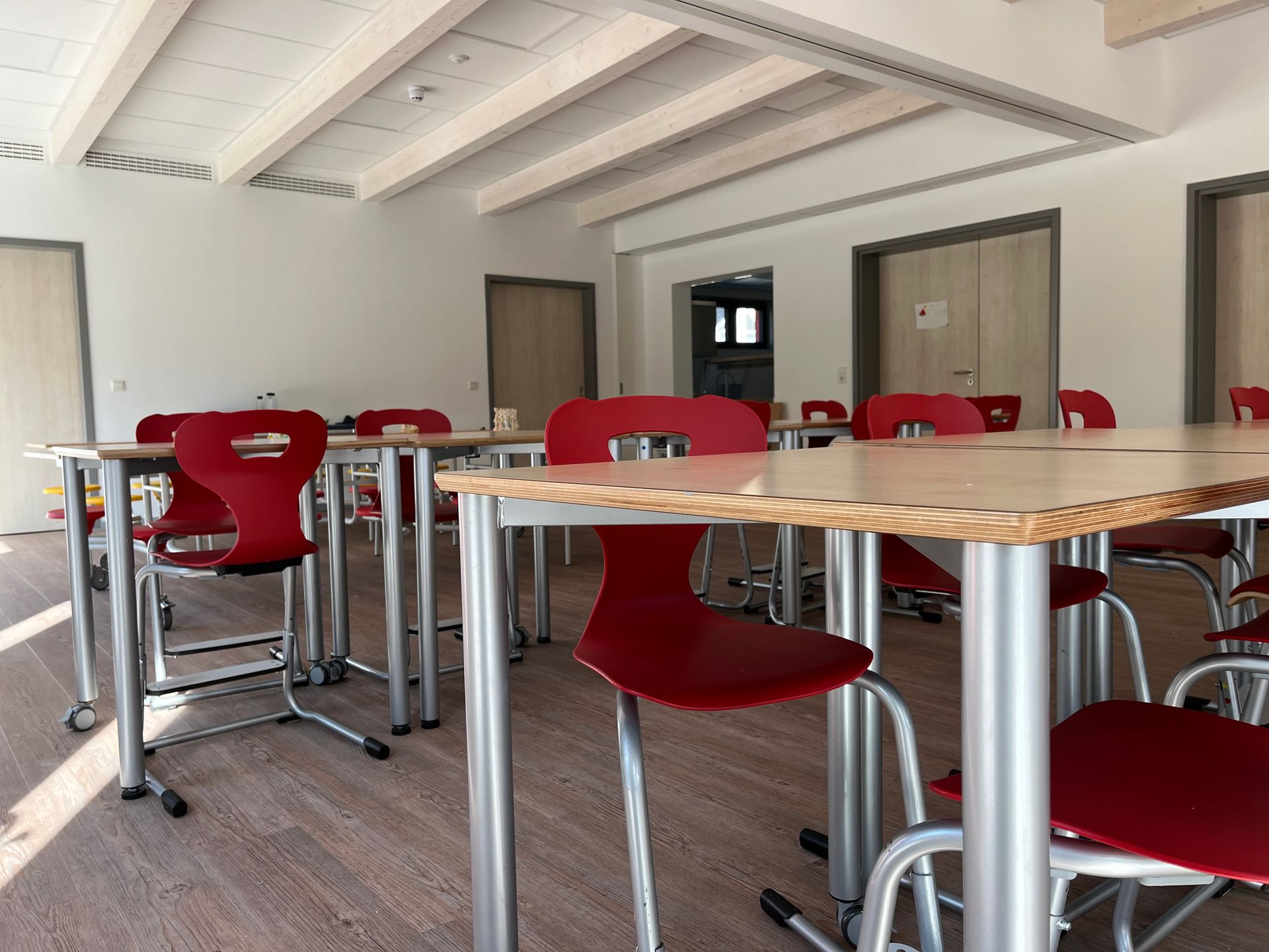 Ein modernes, leeres Klassenzimmer mit Reihen von Holztischen und roten Plastikstühlen auf einem hellen Holzboden.