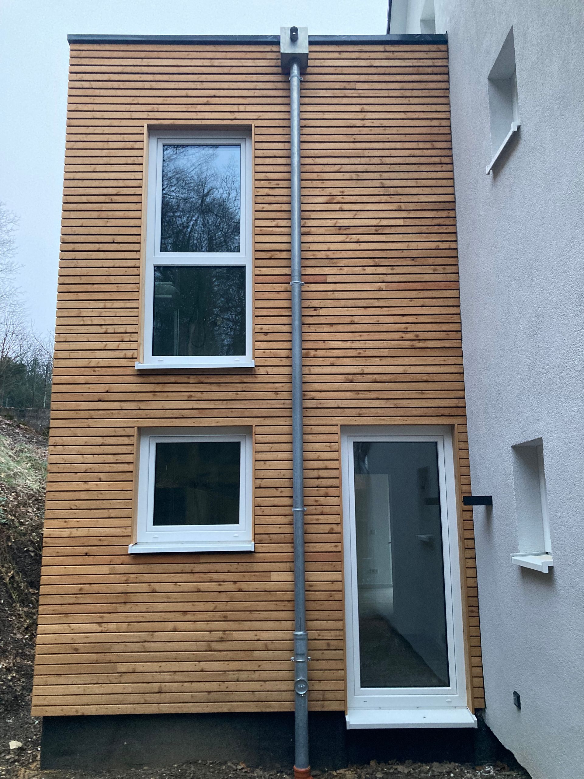 Außenansicht eines modernen Hausanbaus mit horizontaler Holzverkleidung, einem Fallrohr aus Metall, 