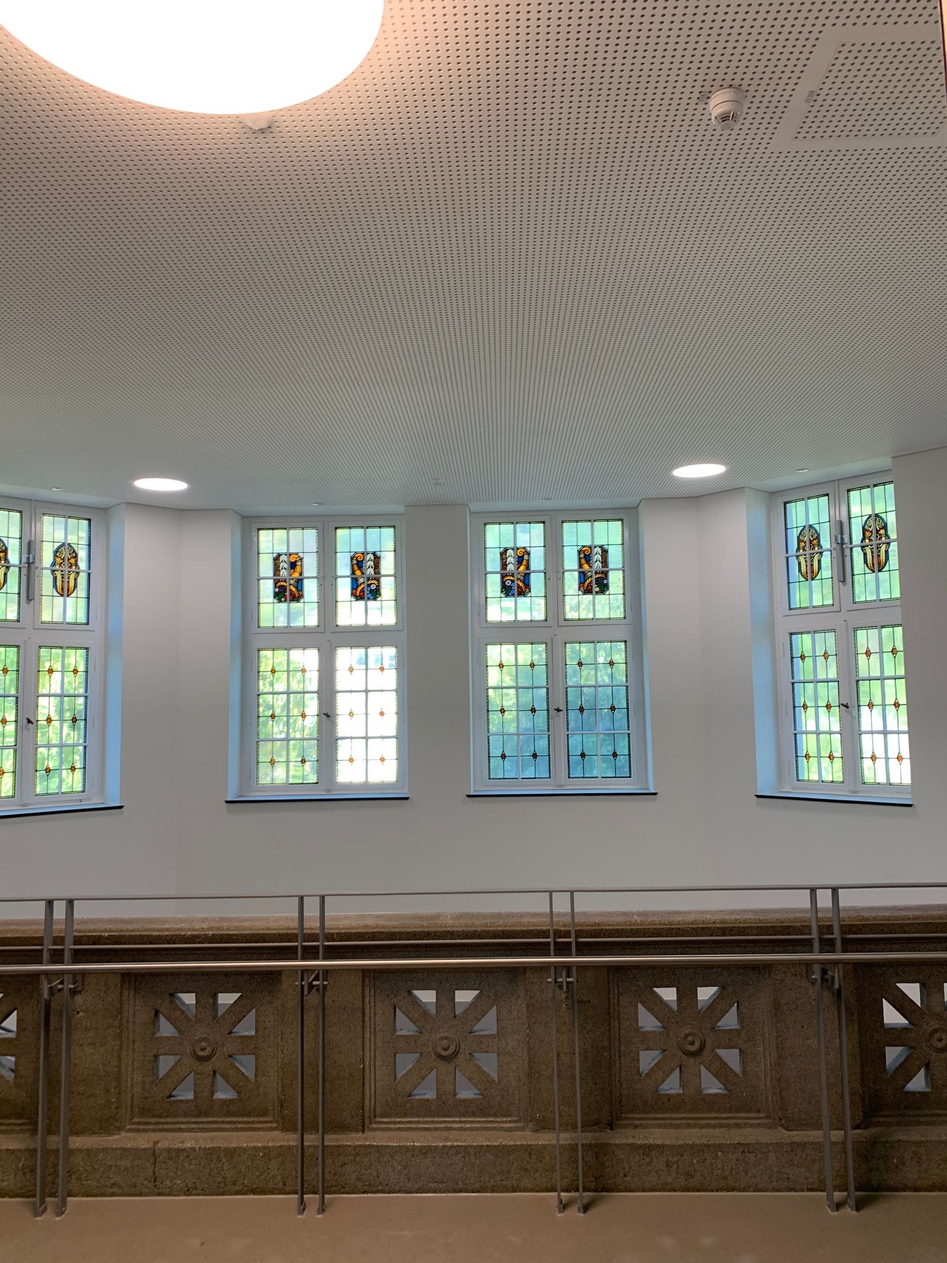 Ein heller Raum mit vier Fenstern mit Buntglas-Wappen, einer gemusterten Akustikdecke und einem steinernen Geländer.
