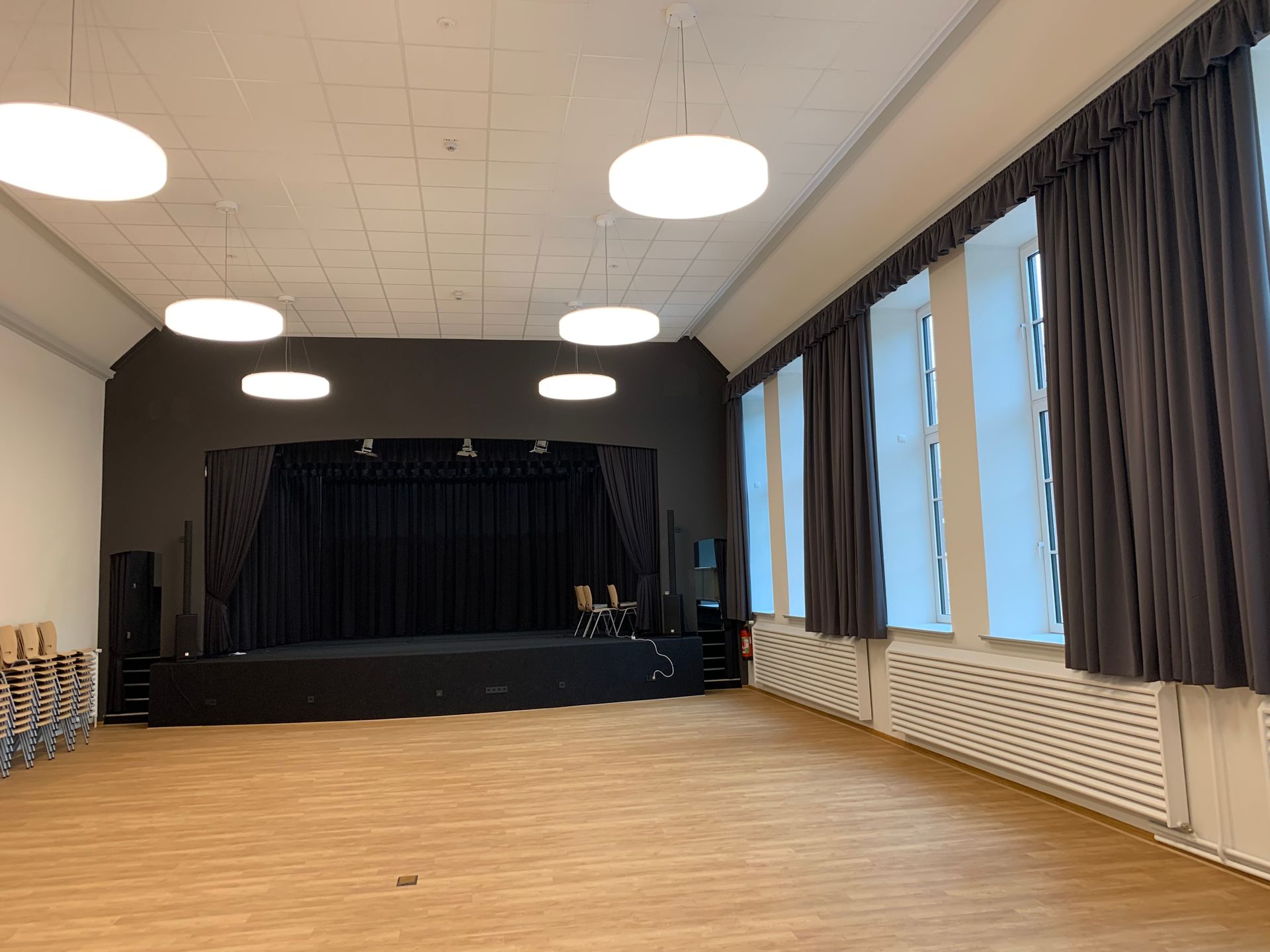 Ein leerer Saal mit Holzboden, eine Bühne mit schwarzen Vorhängen, weiße Deckenleuchten und Fenster mit grauen Vorhängen.