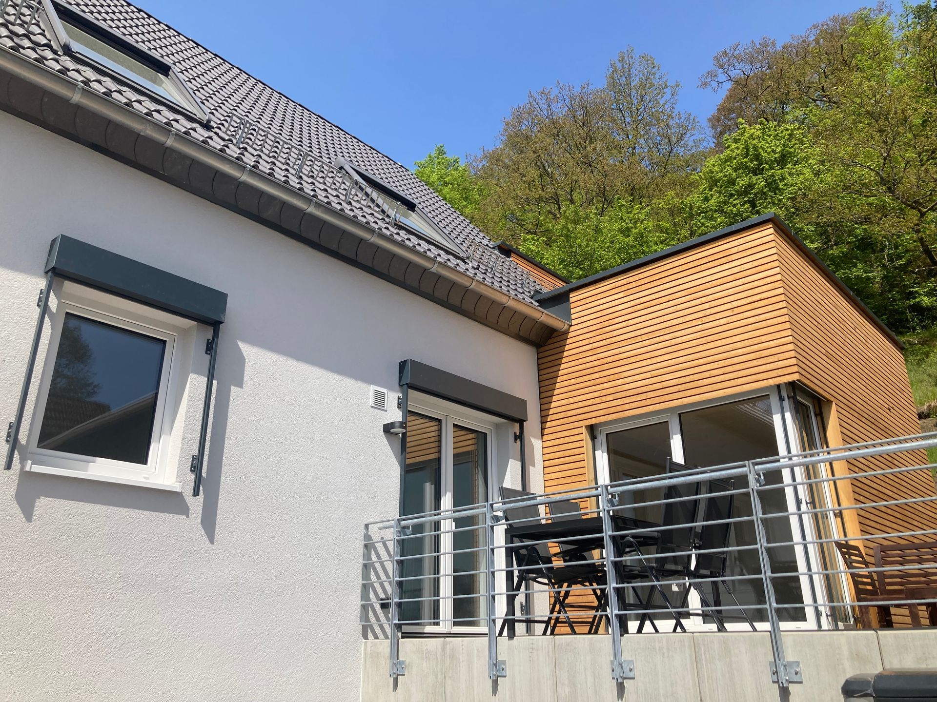 Weißes Haus mit einem modernen, braun gefliesten Anbau, Metallbalkongeländer und Gartenmöbeln unter sonnigem, blauem Himmel.