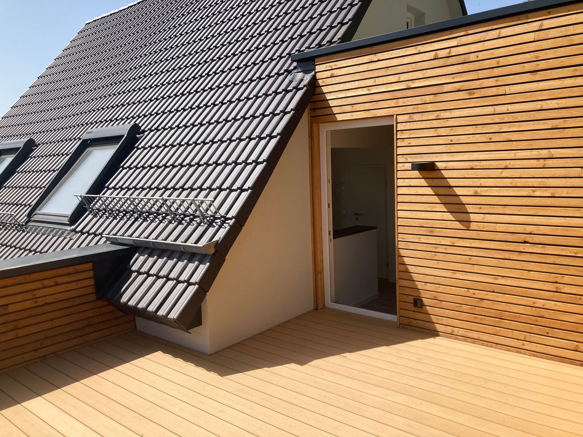 Eine Dachterrasse mit hellen Holzplanken, einem Ziegeldach und einer holzgetäfelten Wand mit offener Tür.