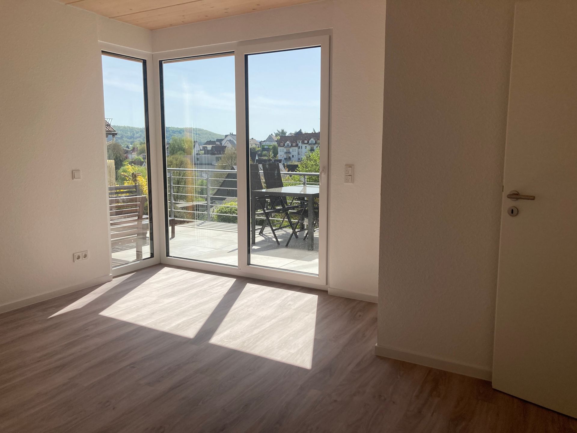 Ein helles, leeres Zimmer mit hellen Holzdielen, weißen Wänden und einem großen Eckfenster, das auf einen Balkon führt.