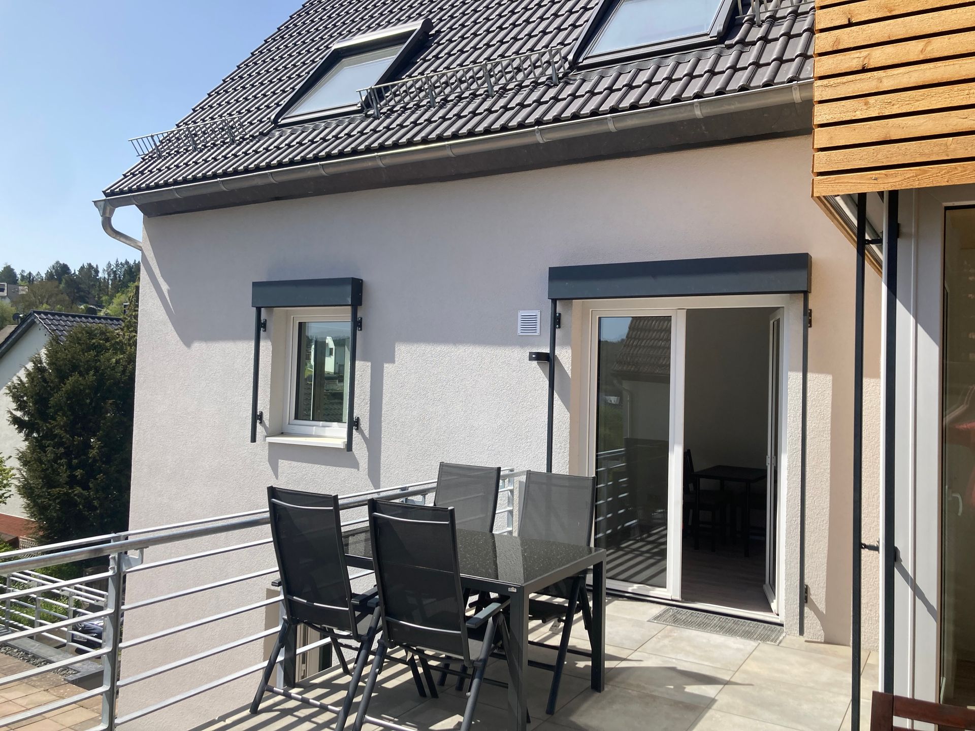 Ein Balkon mit einem dunklen Tisch und vier Stühlen vor einem hellen Haus mit zwei Dachfenstern.