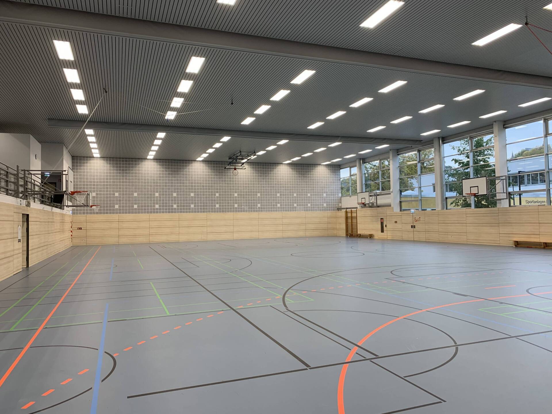 Eine leere Turnhalle mit grauem Boden, Holzwänden, Basketballkörben und einer gemusterten Decke mit Beleuchtung.