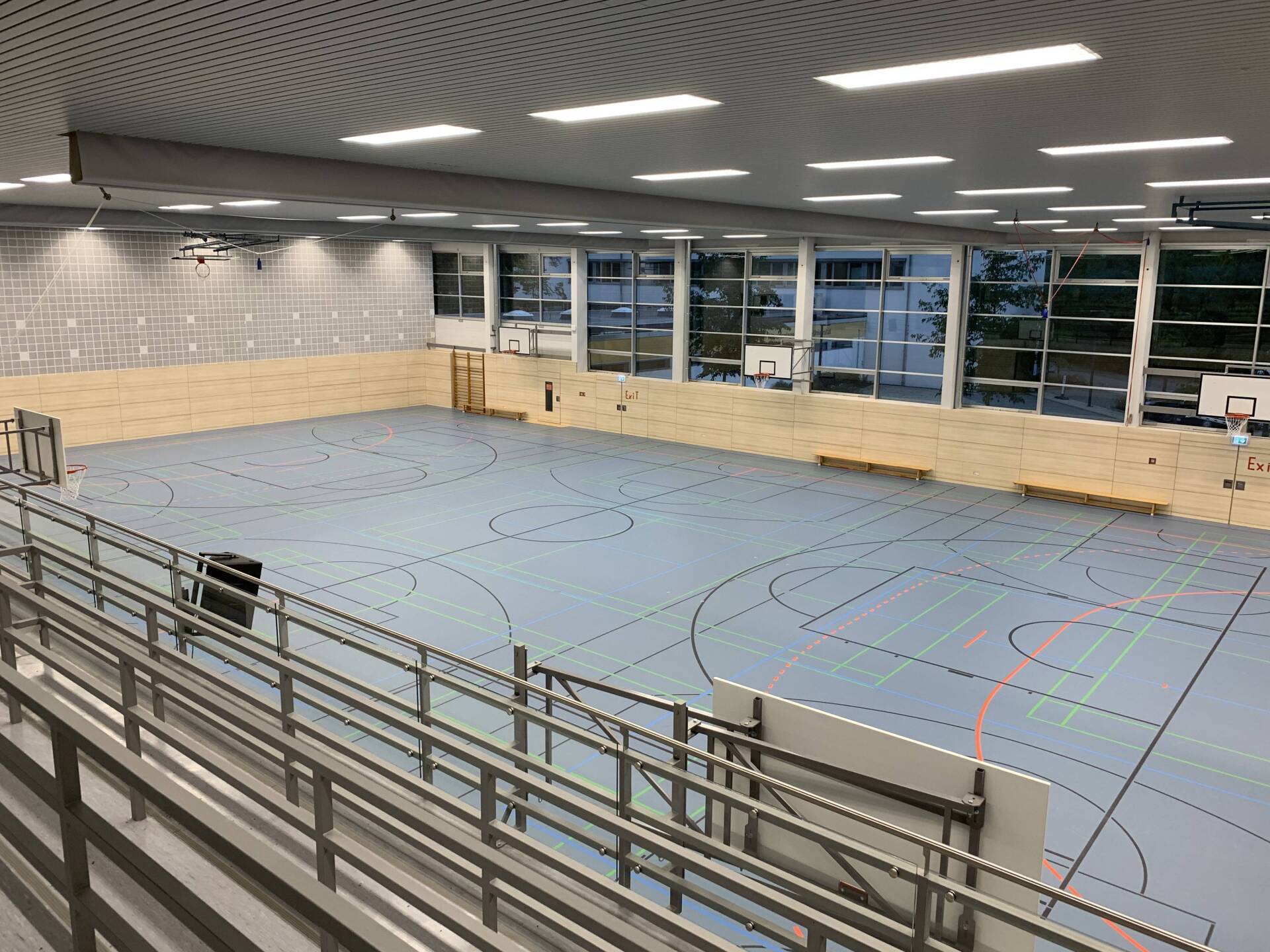 Eine leere Sporthalle mit hellblauem Boden, markierten Spielfeldern, gestuften Sitzreihen im Vordergrund und großen Fenstern.