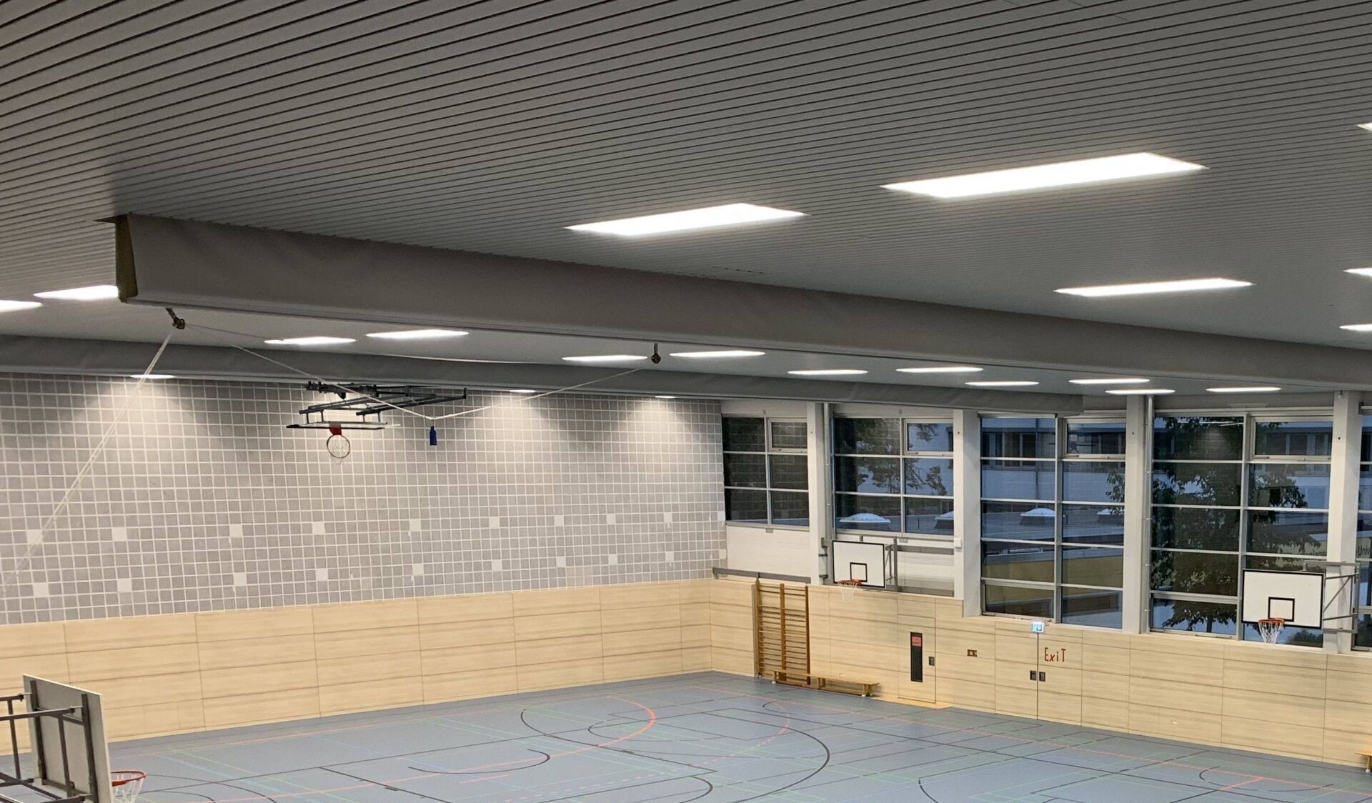 Eine Turnhalle mit hellen Holzwänden, graublauem Boden und einer gemusterten Decke mit rechteckigen Leuchten.