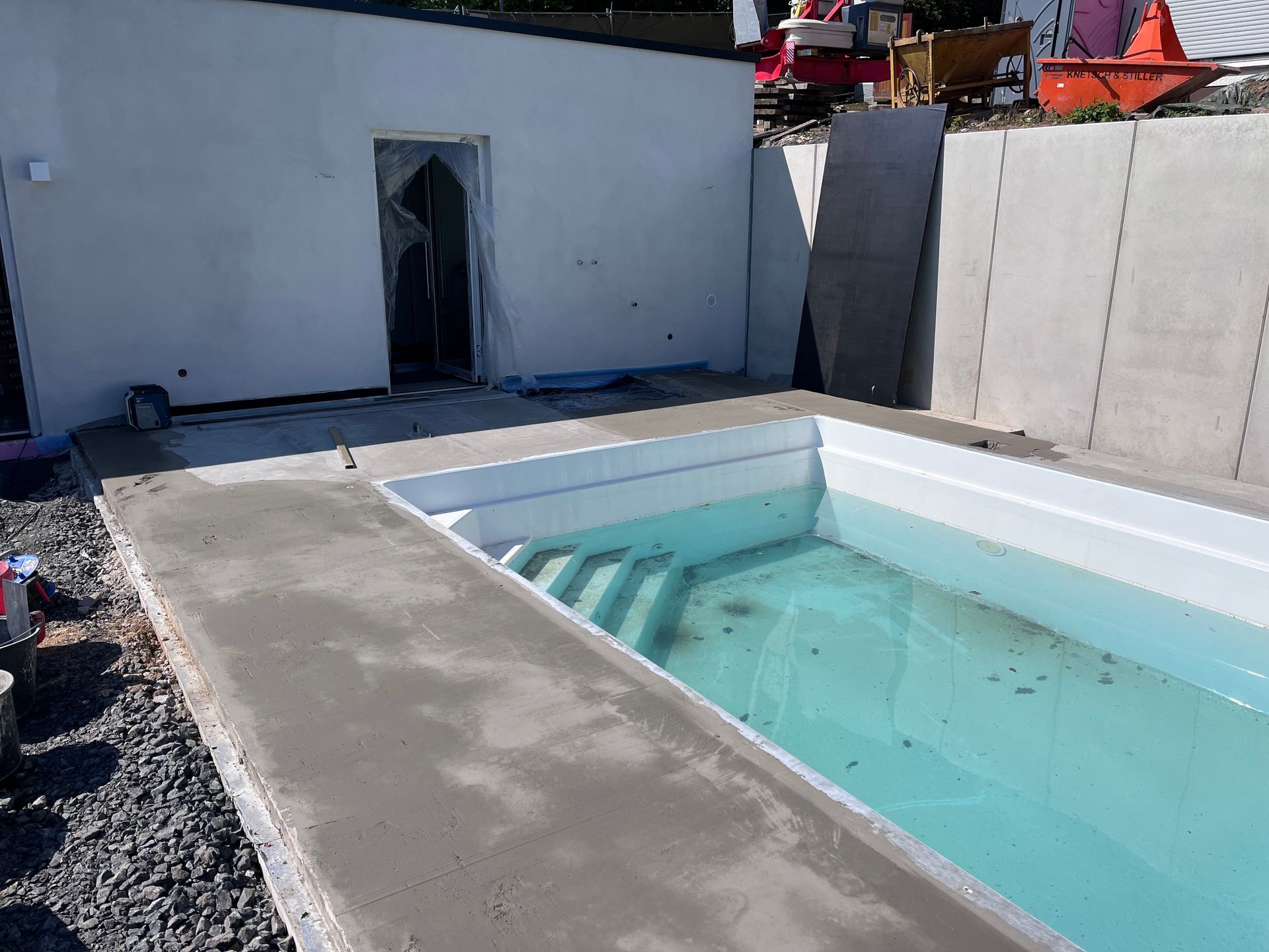Ein im Bau befindliches Schwimmbecken mit nasser Betonplatte, angrenzend an eine weiße Mauer und eine Betonstützmauer.