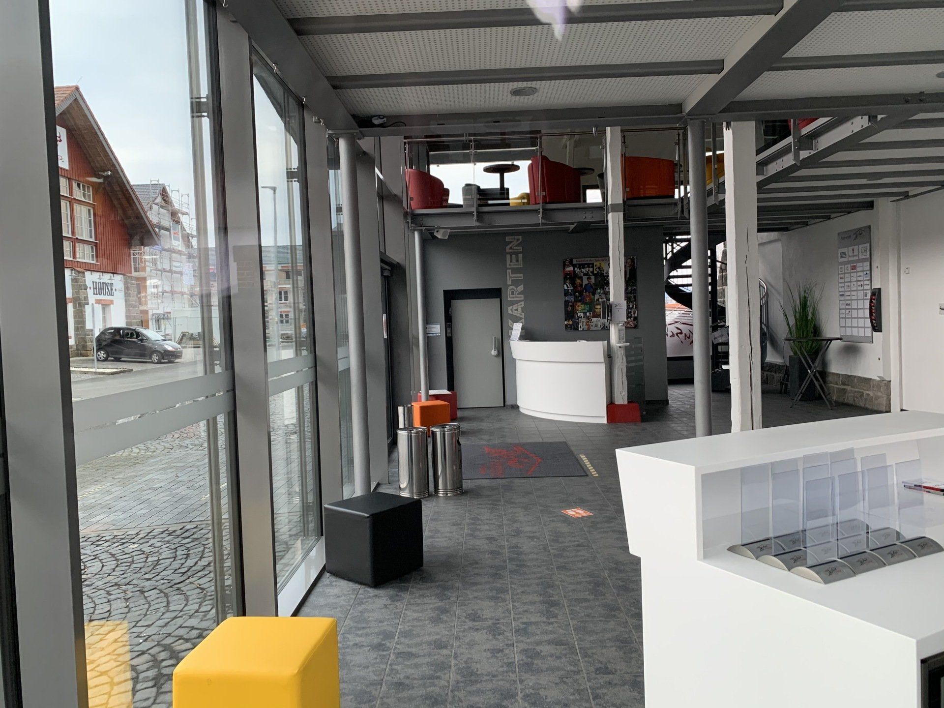 Moderne Lobby mit großen Fenstern, farbenfrohen Sitzgruppen, einem zentralen Empfangstresen und einer oberen Zwischenebene.