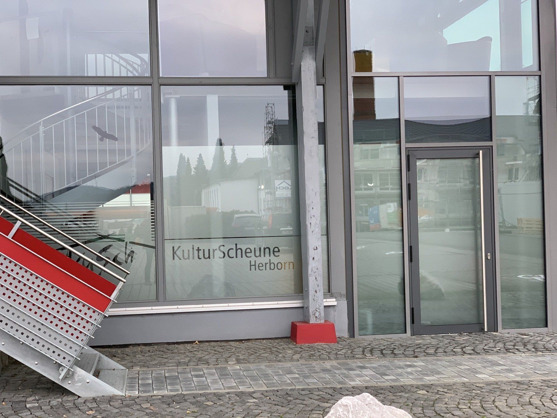 Ein Gebäudeeingang mit Glasfront, rotem Treppenhaus und einem Schild mit der Aufschrift „KulturScheune Herzo“ am Fenster.