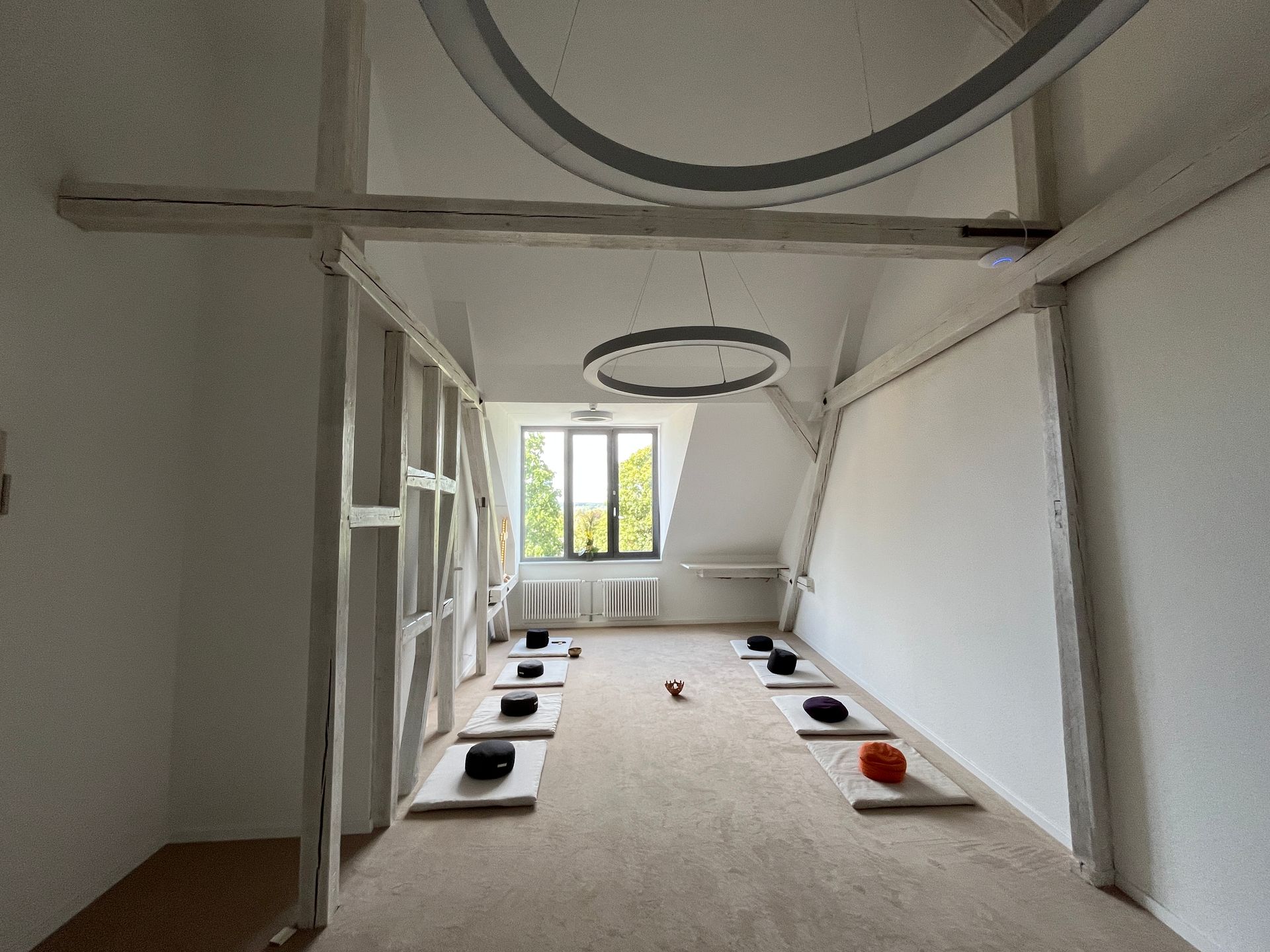 Ein heller Meditationsraum im Dachgeschoss mit weißen Wänden, freiliegenden Holzbalken und zwei Reihen 