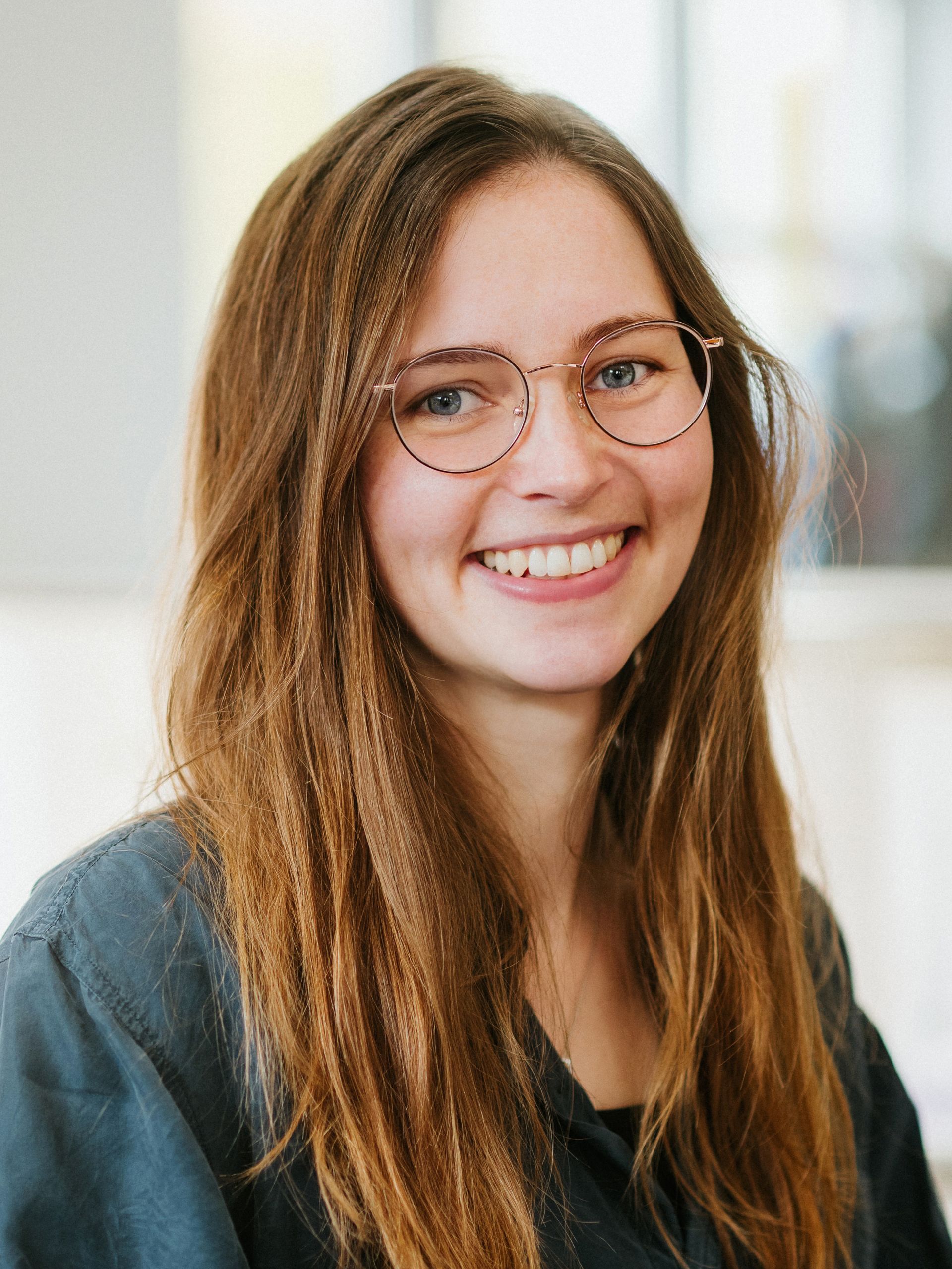 Eine Person mit langen, gewellten braunen Haaren und Brille lächelt warmherzig in die Kamera.