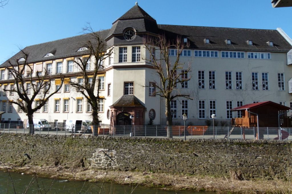 Hinter einem grasbewachsenen Damm und einem Bach steht ein großes, helles, mehrstöckiges Gebäude mit einem Uhrturm